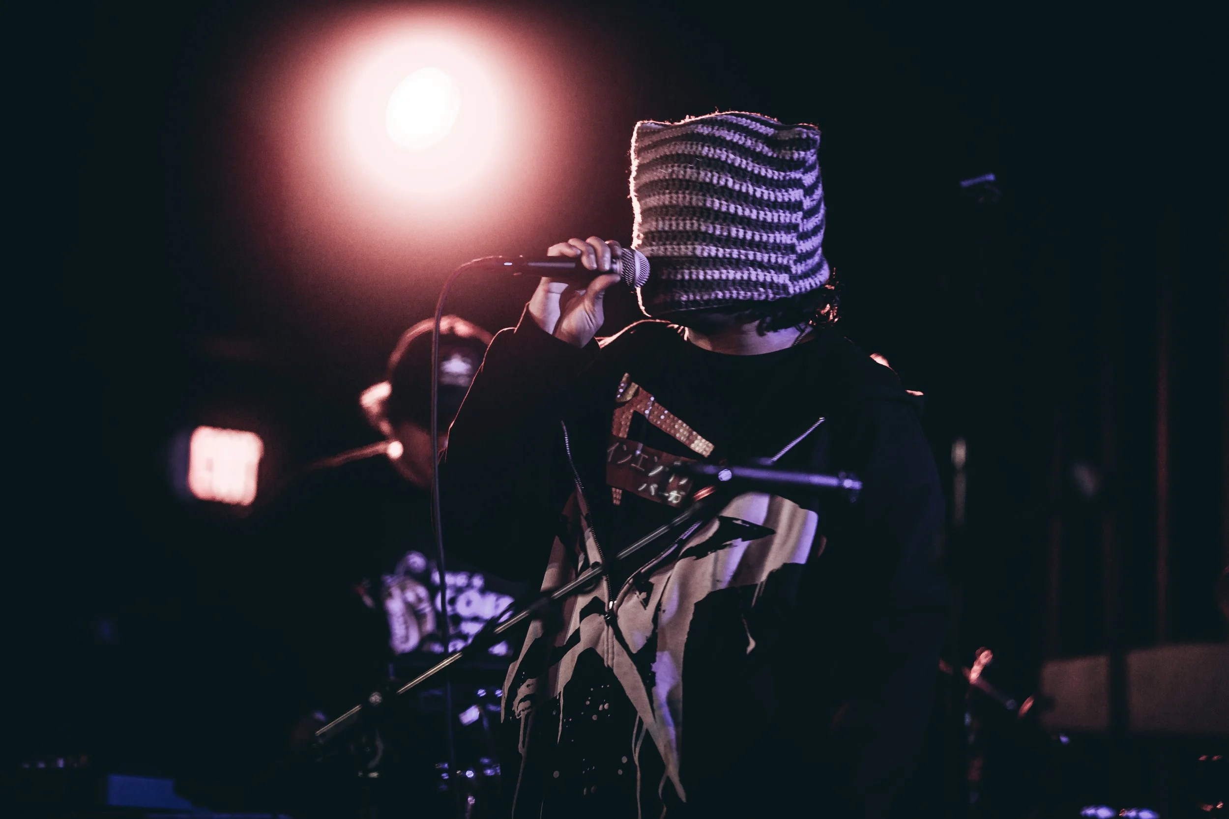 Person wearing a striped beanie and dark clothing holding a microphone on stage with colorful lighting.