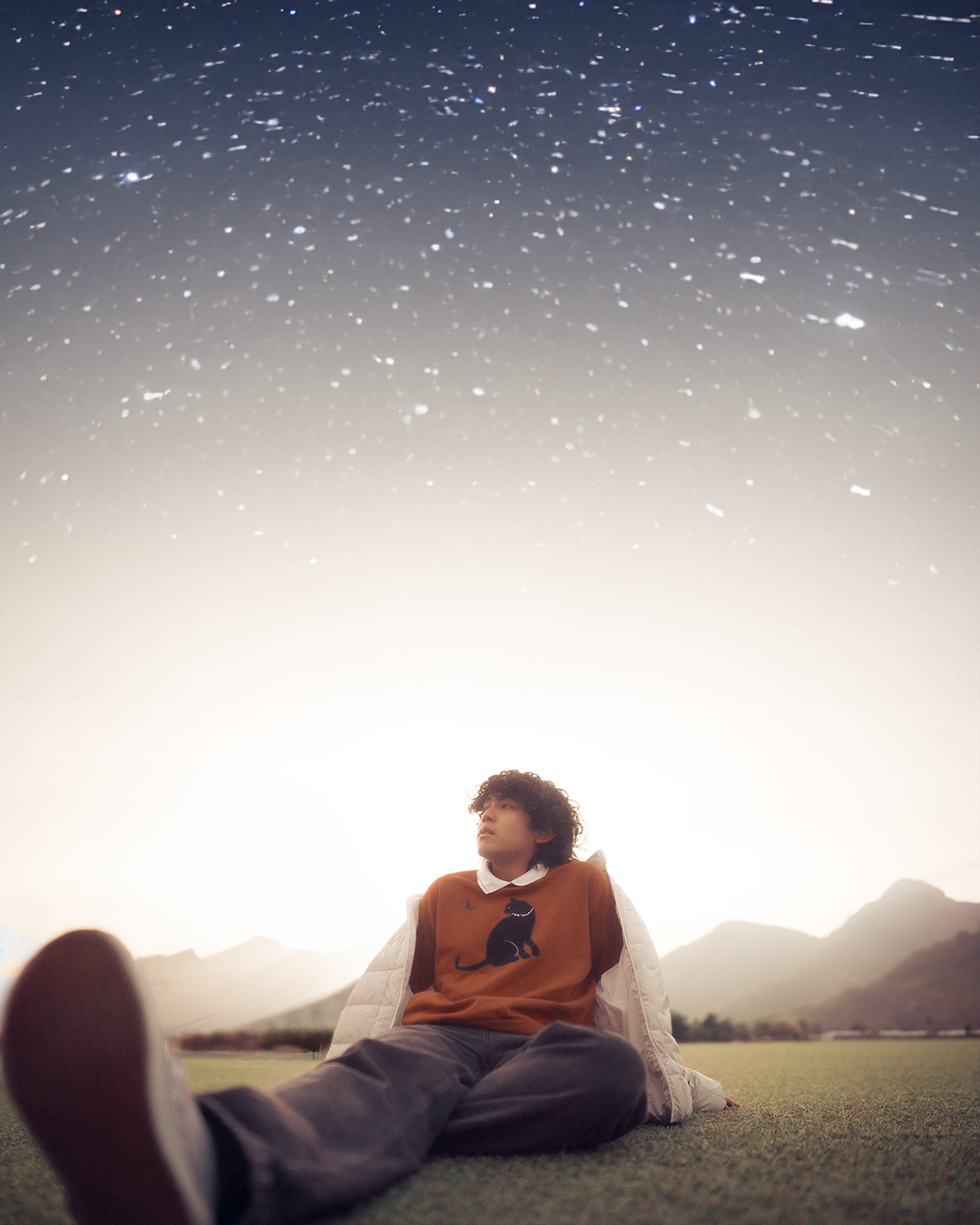 A young man is sitting outdoors on grass, looking up at a night sky filled with stars and galaxy light, with mountains in the background.