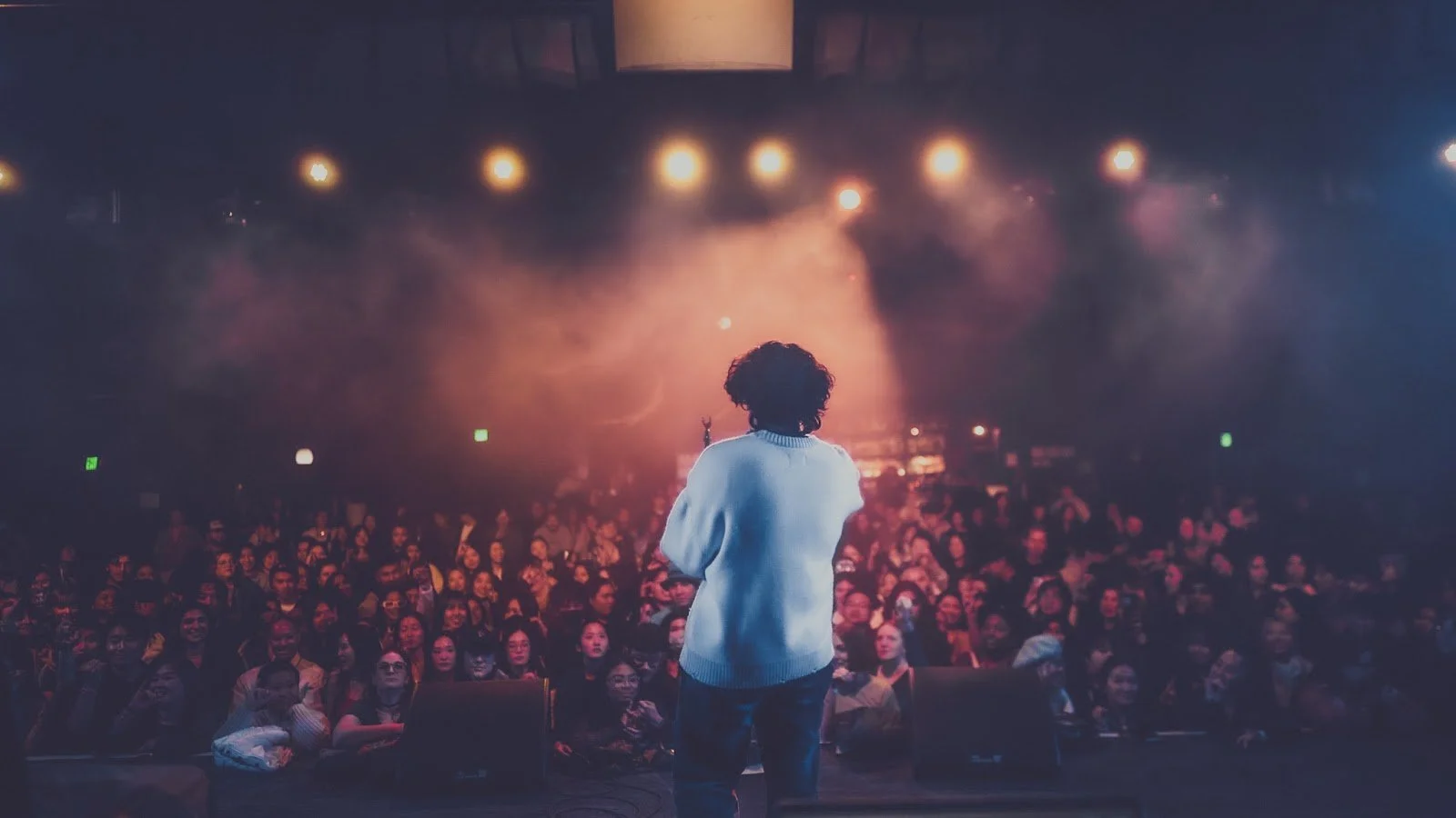 A person with curly hair and a white sweater stands on stage facing a large, smiling audience in a dimly lit venue with stage lights and fog.