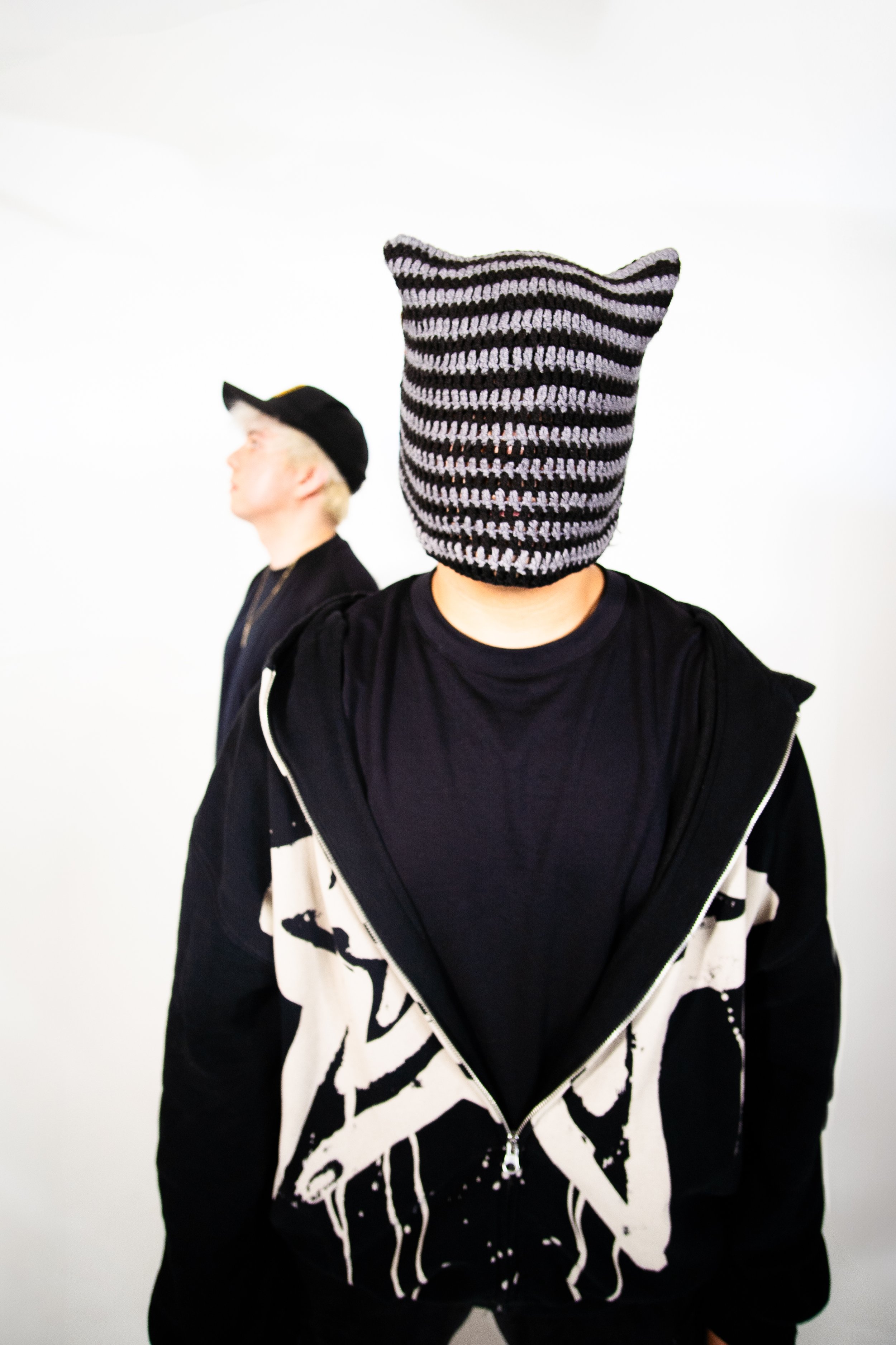Person wearing a black hoodie with white graffiti design, a black shirt underneath, and a colorful striped knit mask resembling a cat's head. The person is standing in front of a plain white background. Another person wearing a black cap and black ja