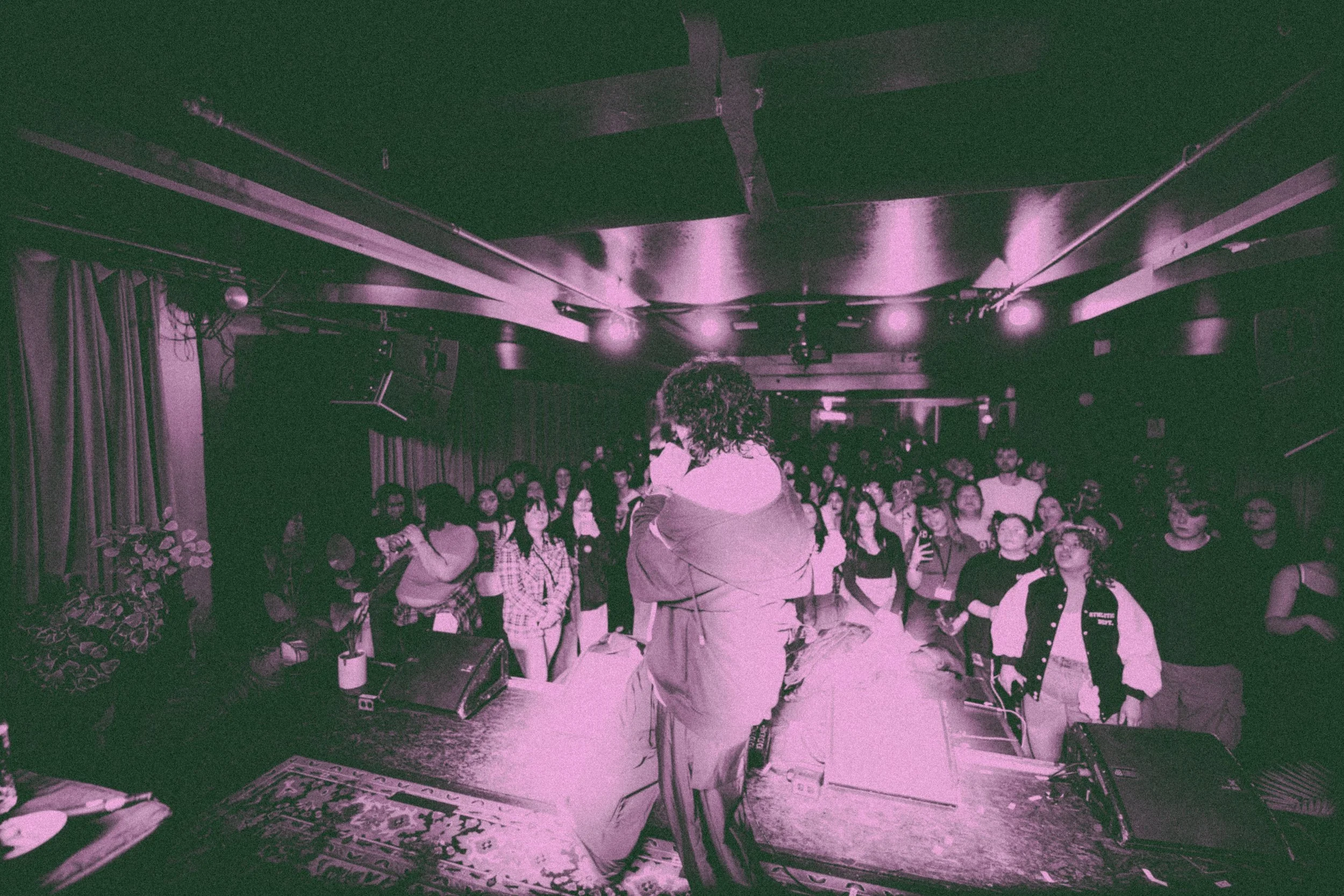 Performer on stage with back facing audience, audience members watching and taking photos, inside a dimly lit venue with ceiling lights.