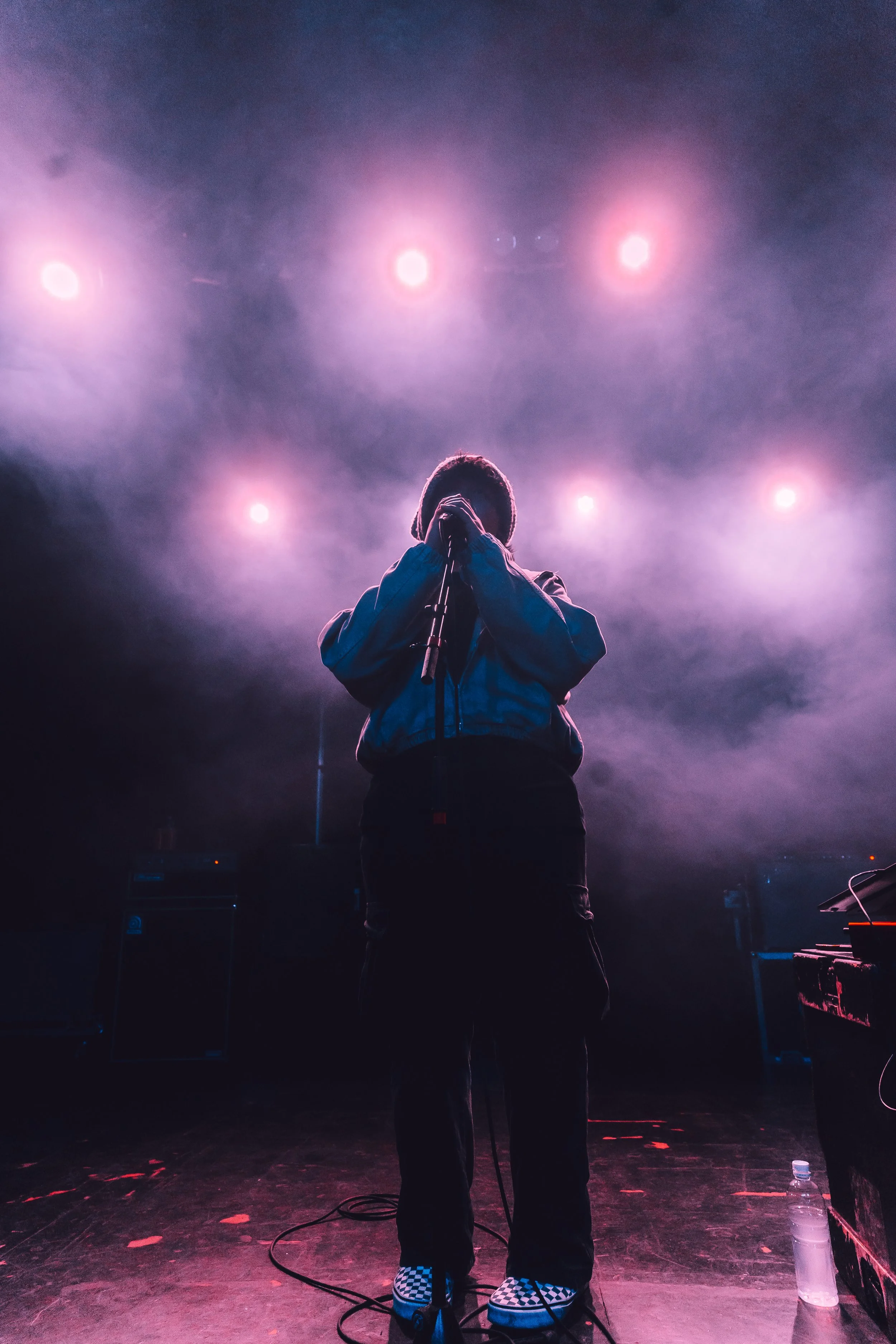 A person standing on stage with purple and pink lights overhead, holding a microphone close to their face. The person is wearing a jacket and checkered shoes, with music equipment and a water bottle nearby.
