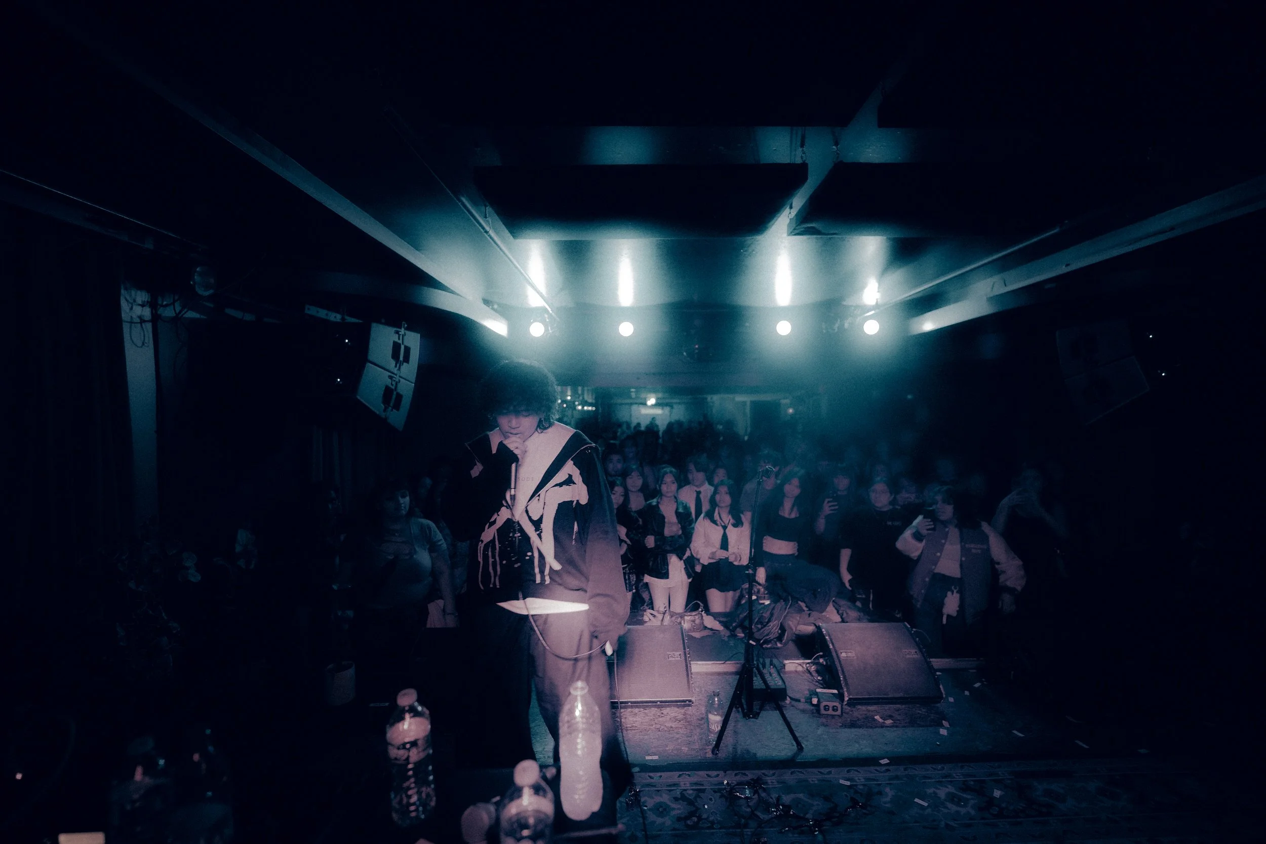 A person standing on stage, hip-hop or rap artist, with a crowd of fans in the background at a concert or performance venue.