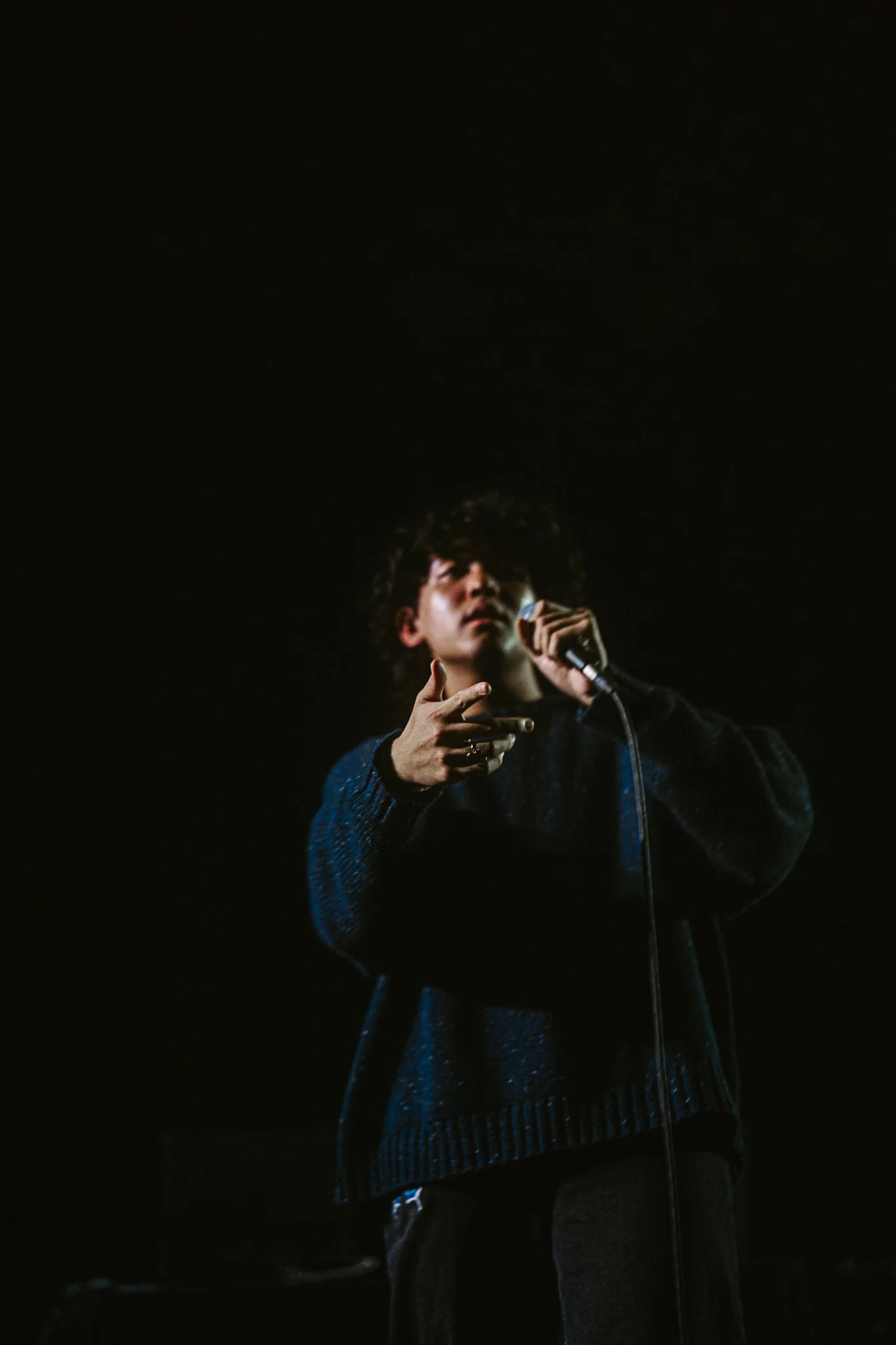 A young man with curly hair performing on stage with a microphone, wearing a dark sweater, against a dark background.