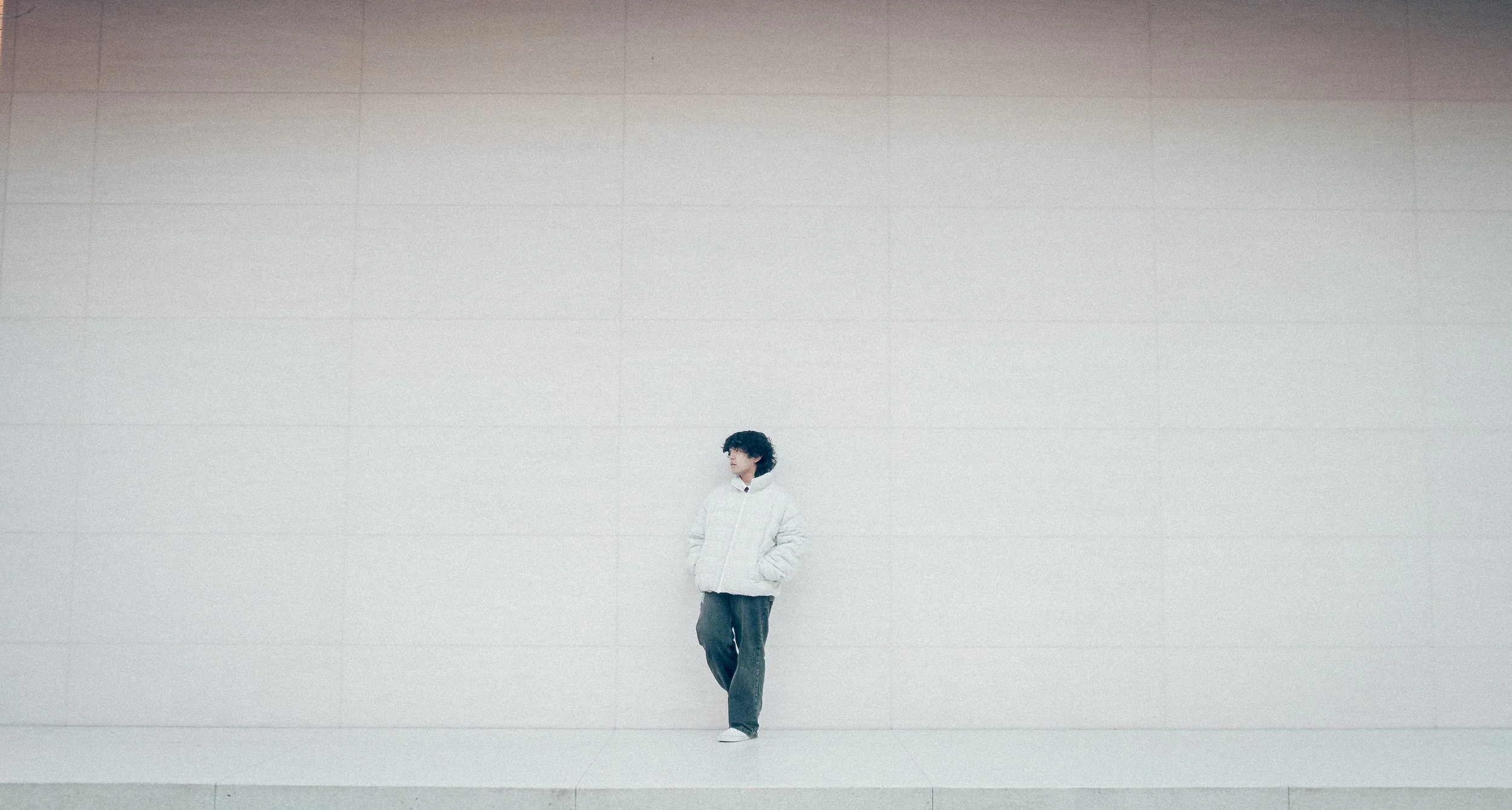 A person with curly hair wearing a white jacket and dark pants leaning against a large, plain, light-colored wall.