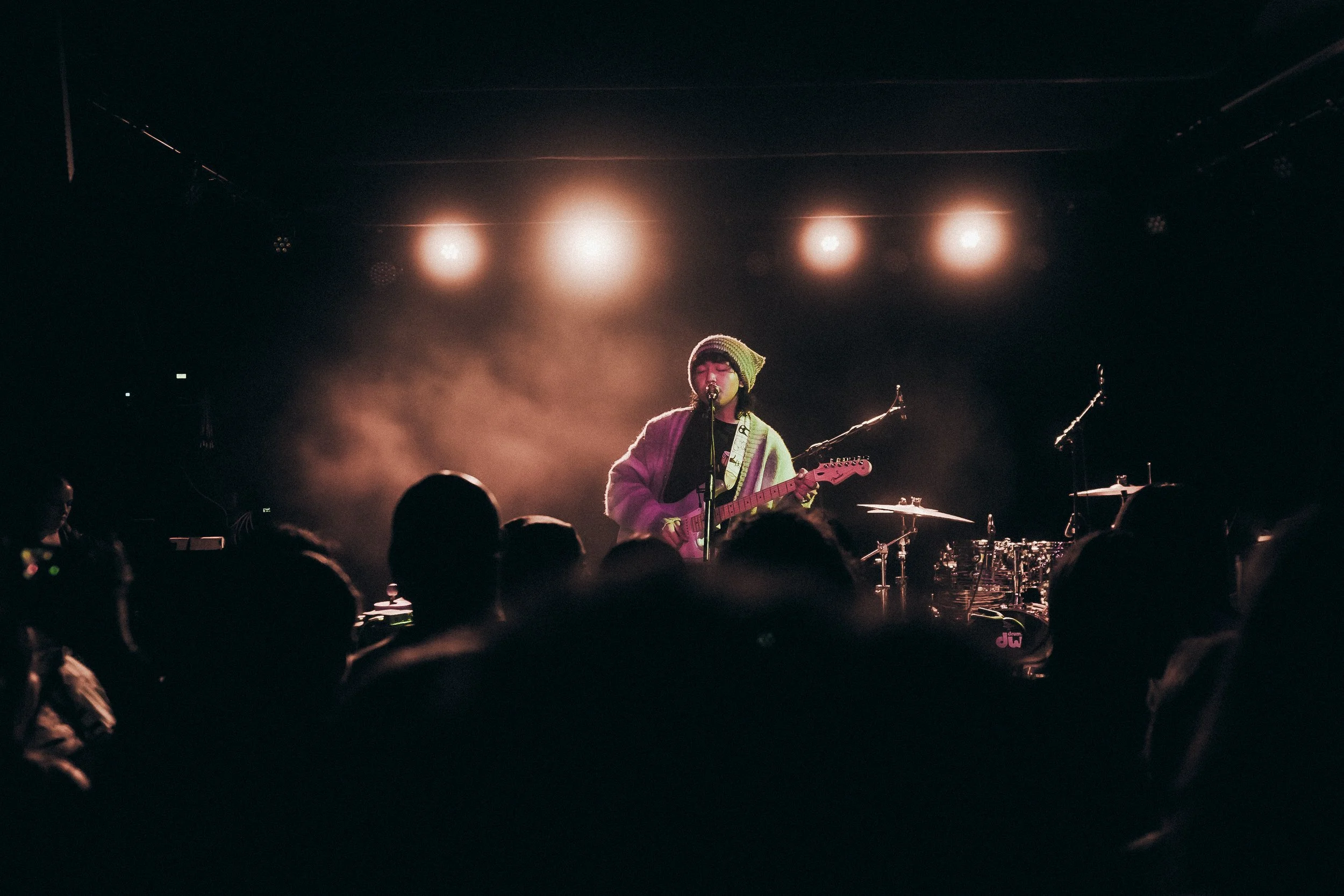 A musician performs on stage with a guitar, wearing a beanie and a cozy sweater, under bright stage lights, with an audience watching.