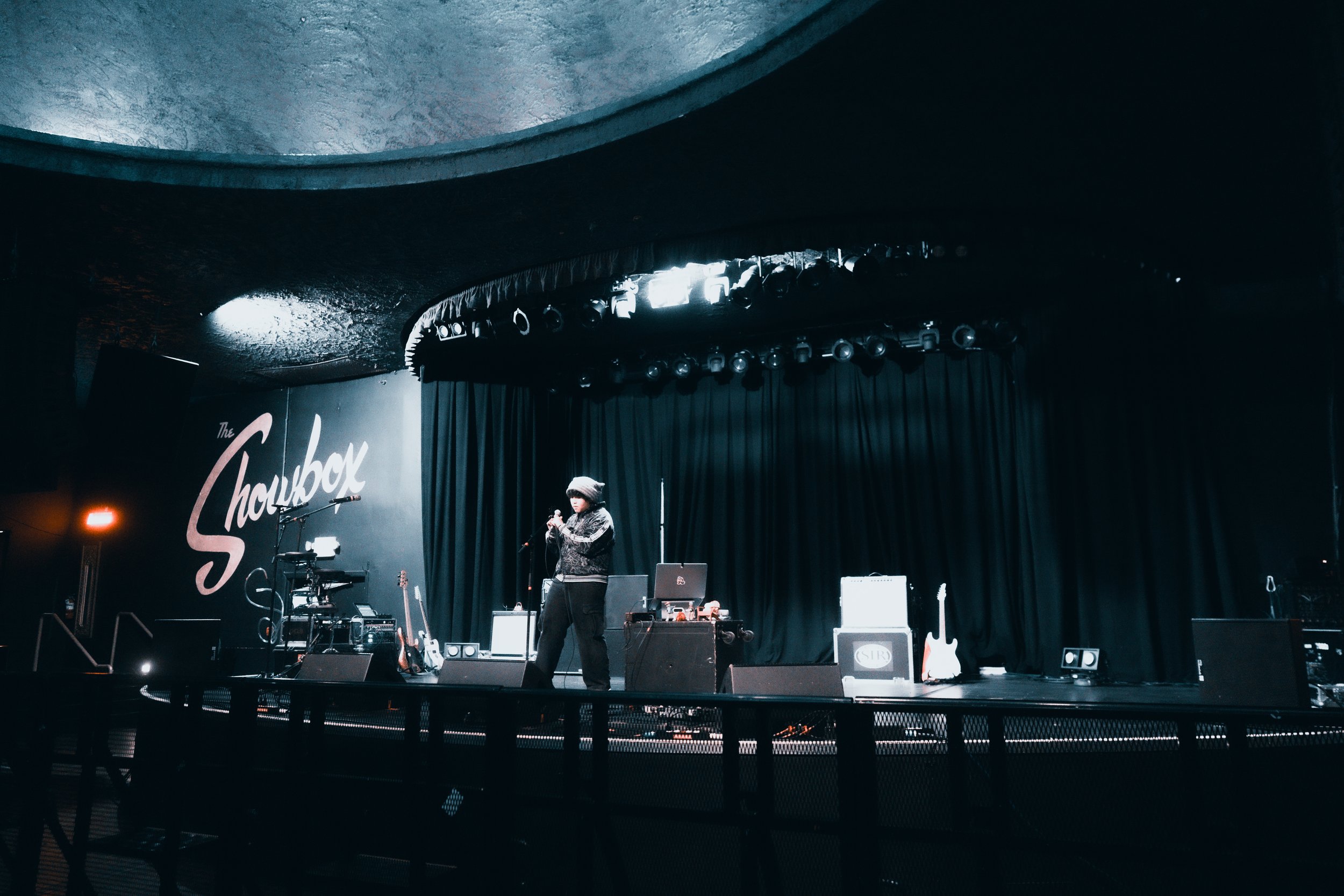 A performer on stage at a music venue called The Chowbox, holding a microphone with musical instruments and equipment around. The stage is dark with a black curtain backdrop and stage lights above.