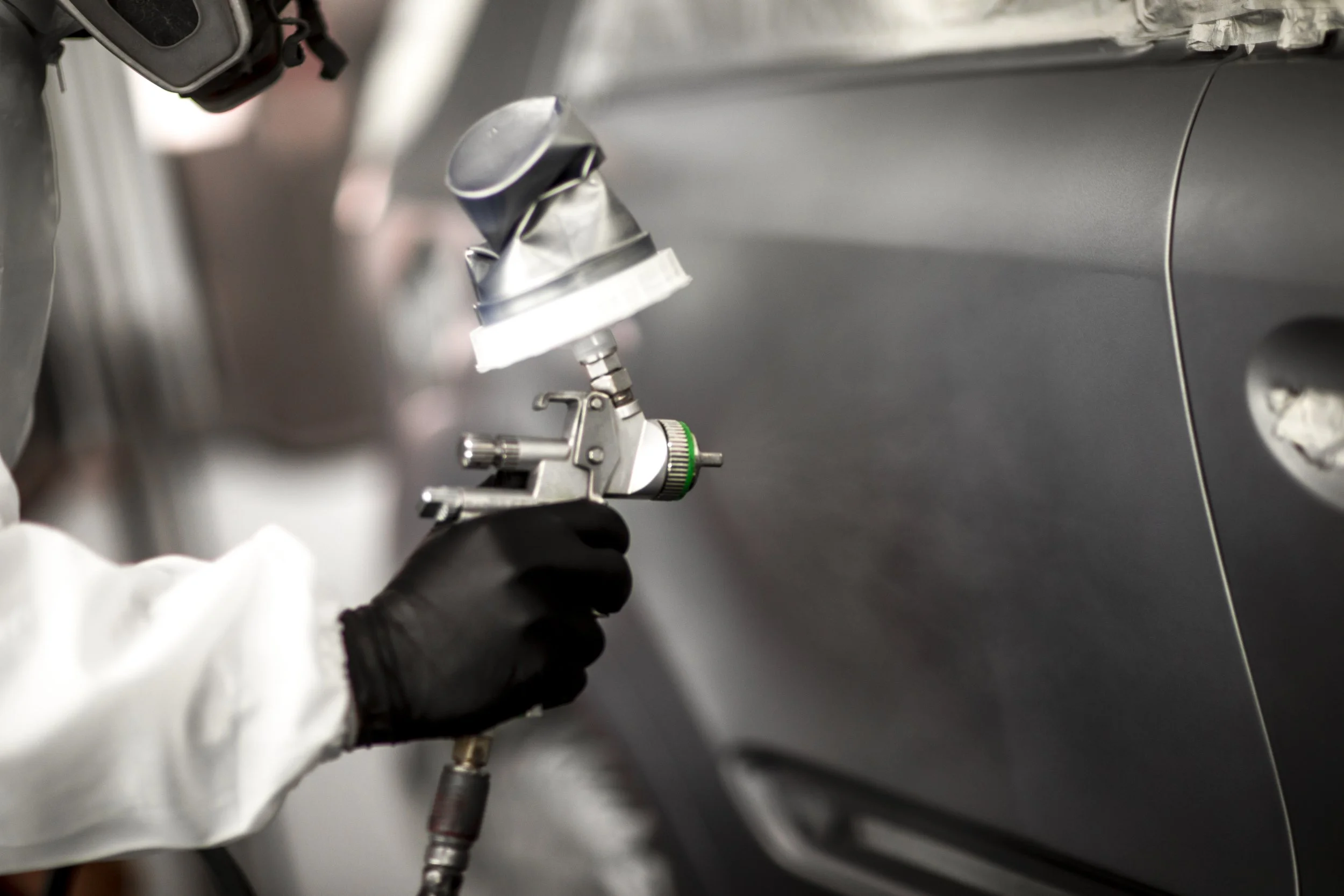 Close-up of a person wearing black gloves and a white lab coat using a spray gun to apply paint or finish to a gray metal surface.