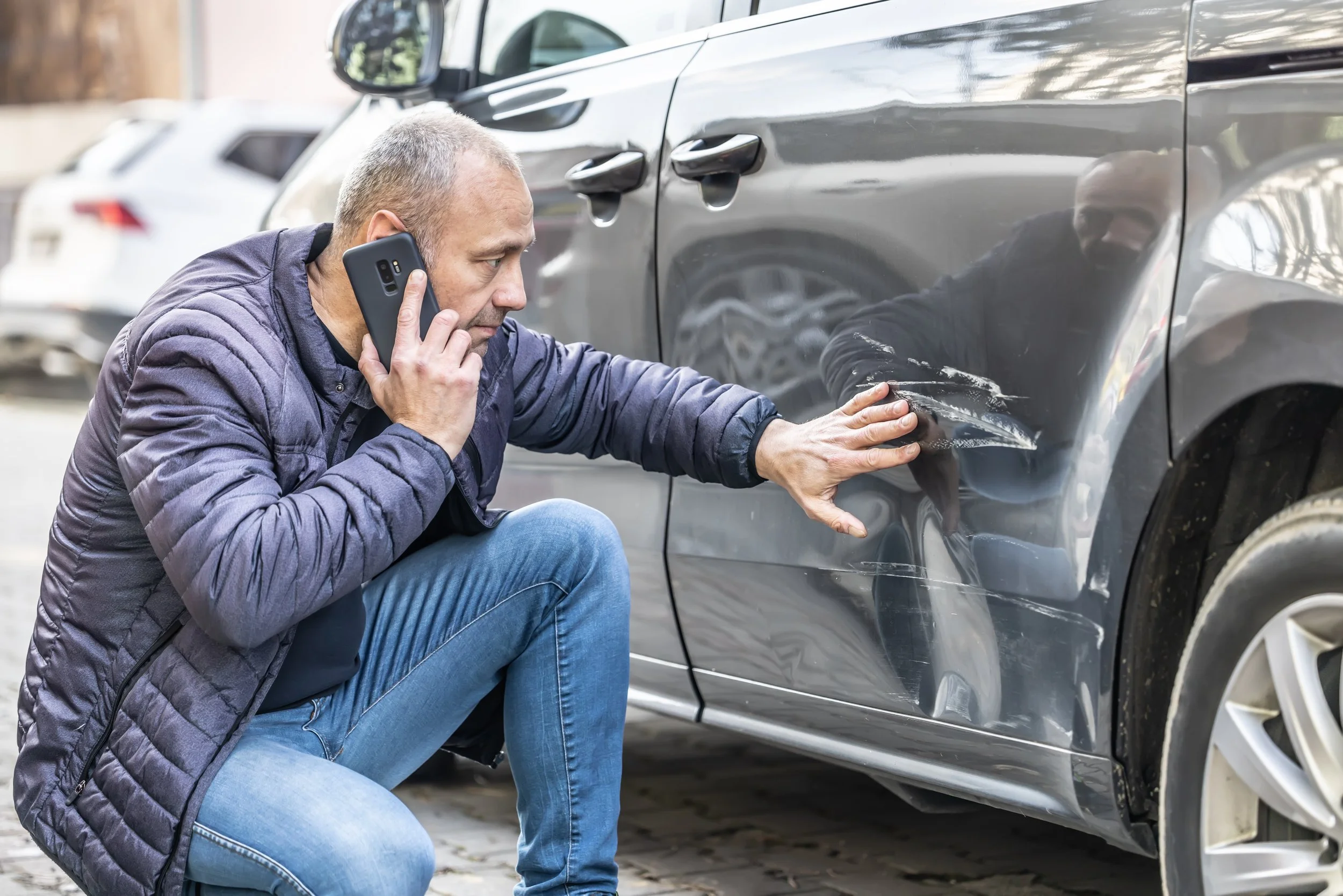 A man with a phone to his ear kneels beside a gray car, examining and touching a scratch or small dent on the car's side panel.