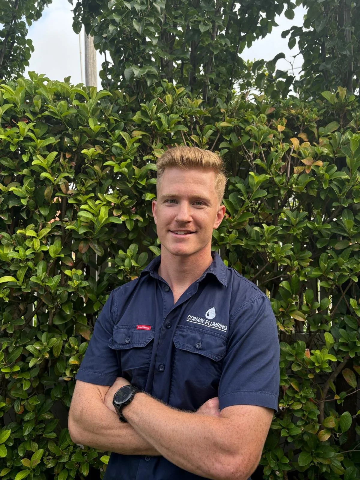 A young man with light skin, blonde hair, and a friendly expression, wearing a navy blue Cormay Plumbing uniform shirt, standing with his arms crossed in front of a lush green shrubbery. He is outdoors under cloudy skies.