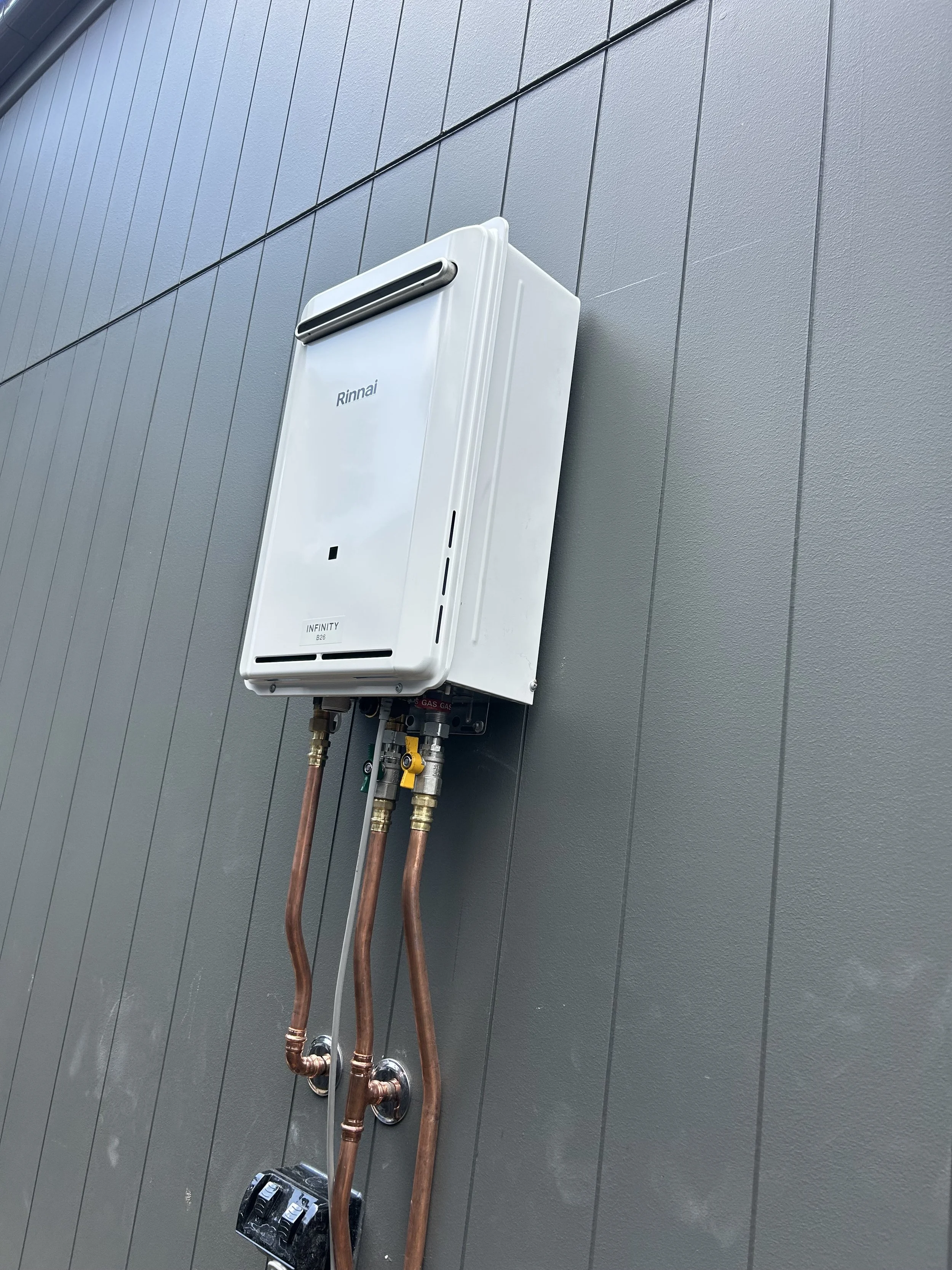 A white Rinnai Infinity tankless water heater mounted on a gray exterior wall with copper pipes connected below.