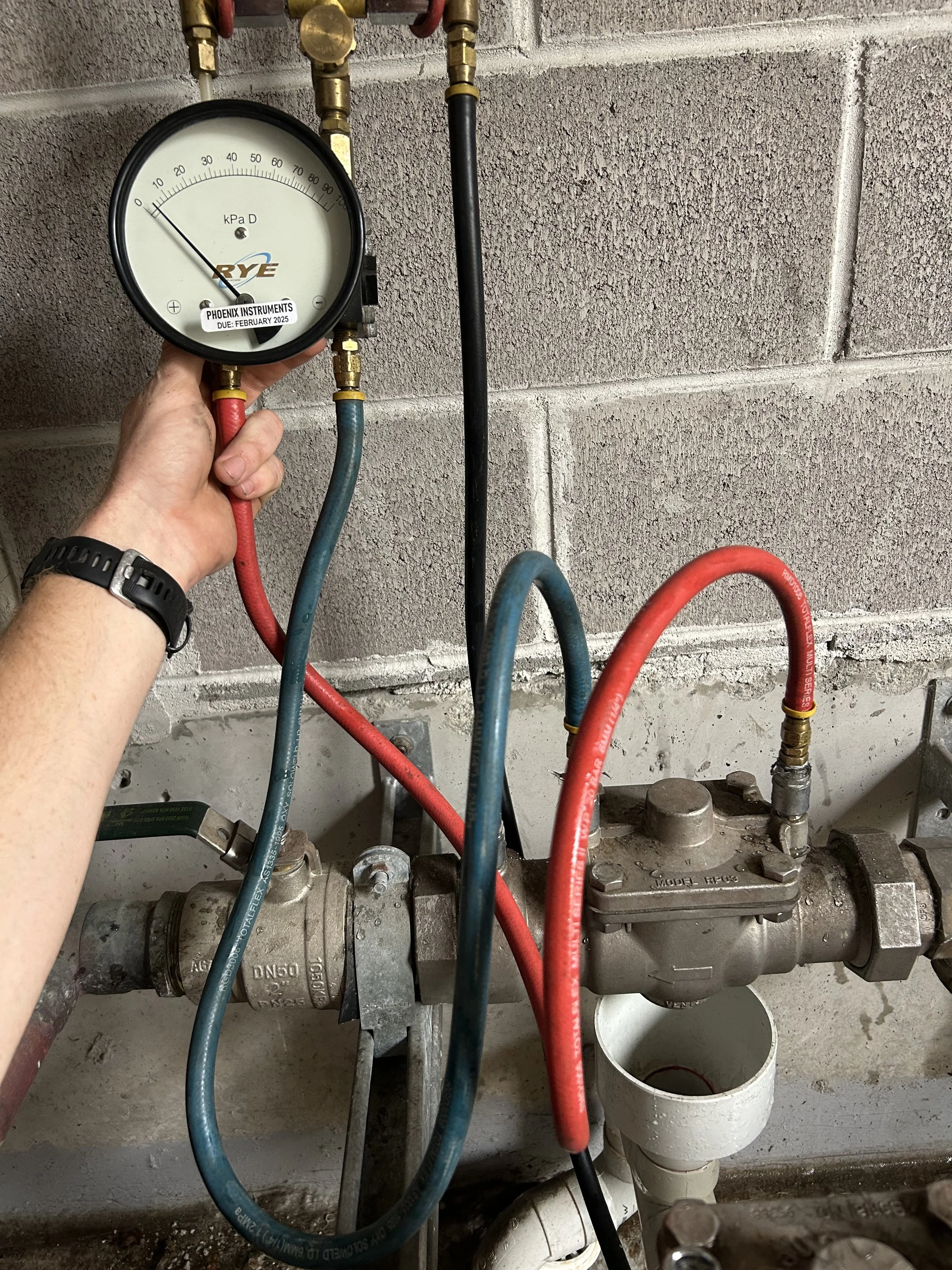 A person's hand holding a pressure gauge connected to plumbing pipes, with a brick wall in the background. The gauge indicates pressure in kilopascals.