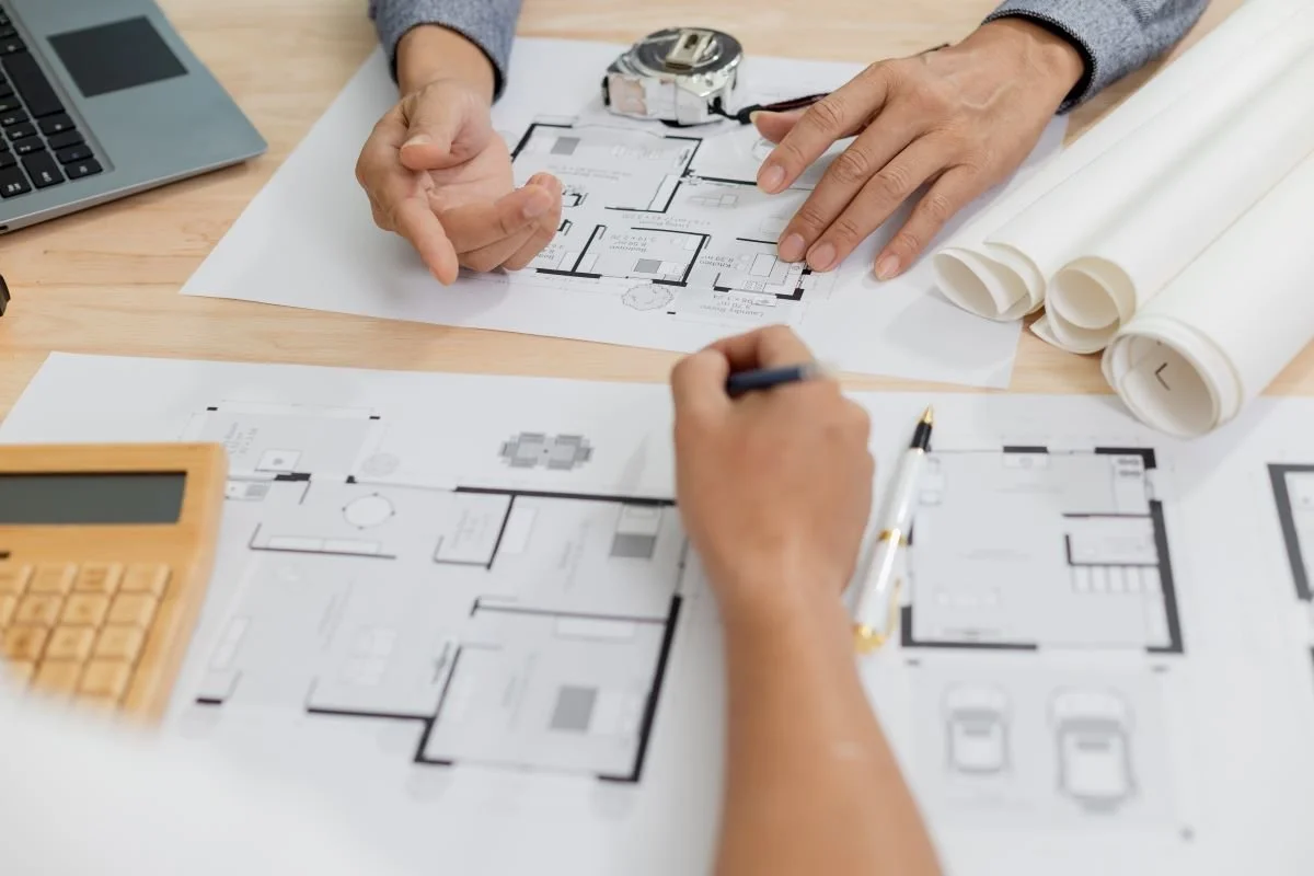 Two people reviewing and discussing architectural blueprints on a wooden table, with a laptop, calculator, tape measure, and rolled-up blueprints nearby.