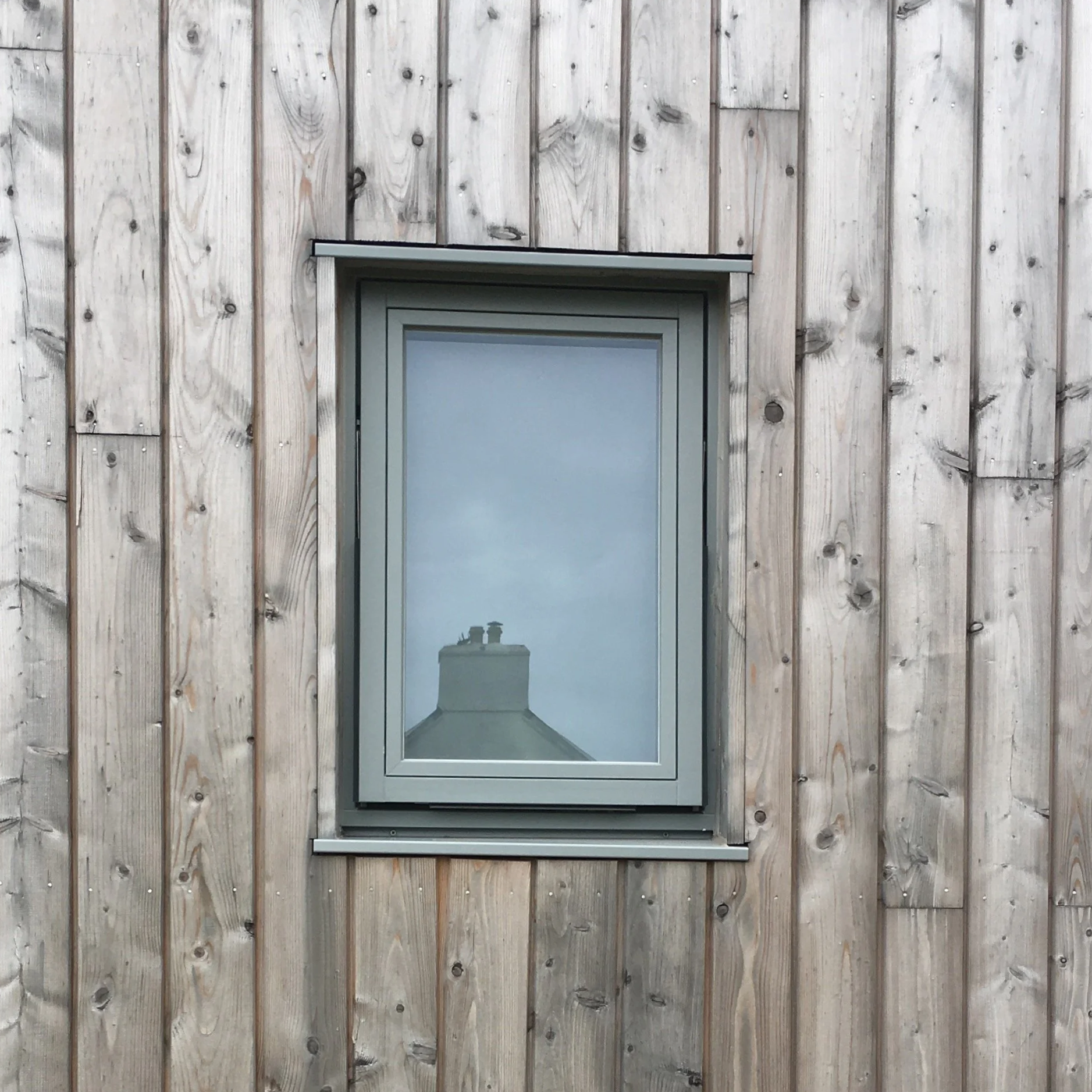 A window on a wooden exterior wall, with the reflection of a chimney with two vents on a gray, overcast sky.