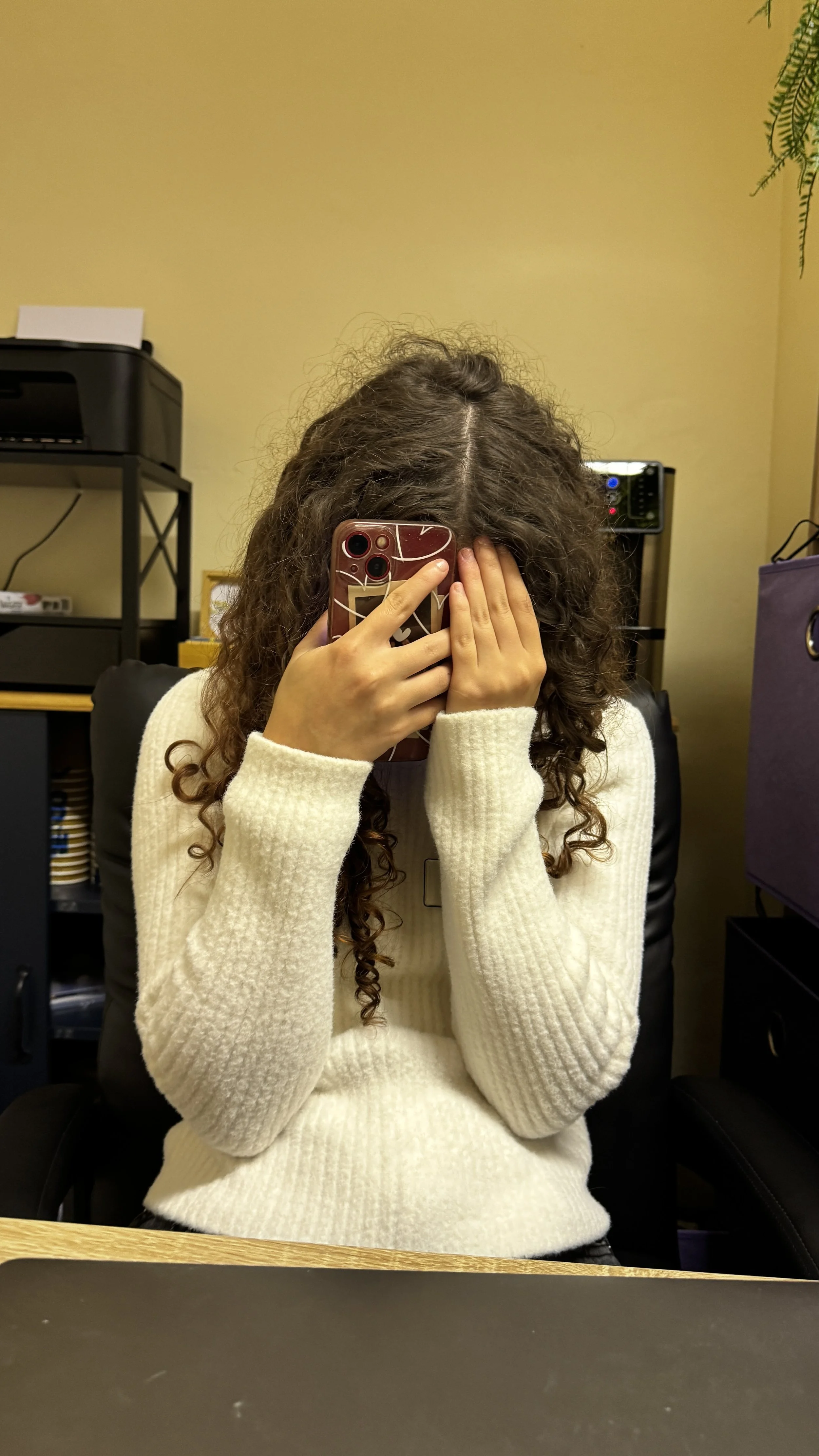 Girl with curly hair taking a mirror selfie, covering half of her face with her phone, seated at a desk in an office setting.