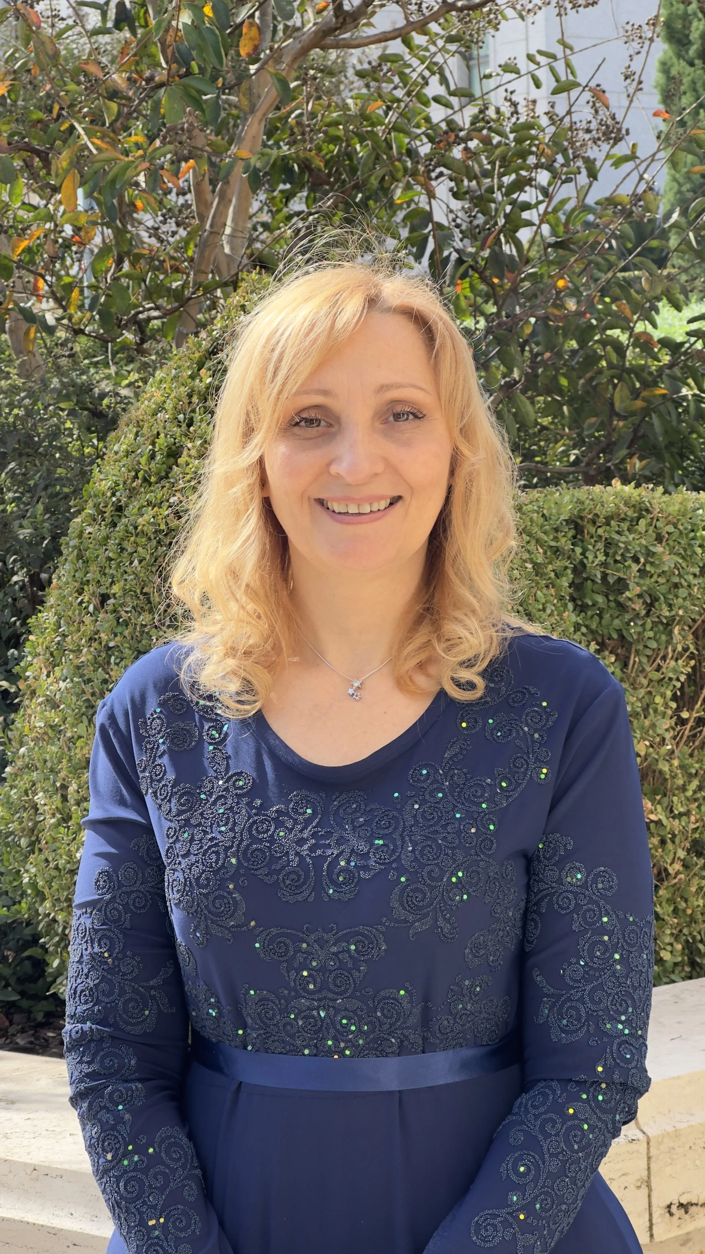 A woman with blonde, curly hair smiling at the camera, wearing a navy blue dress with intricate embroidery and small embellishments, standing outdoors in front of green bushes and trees.