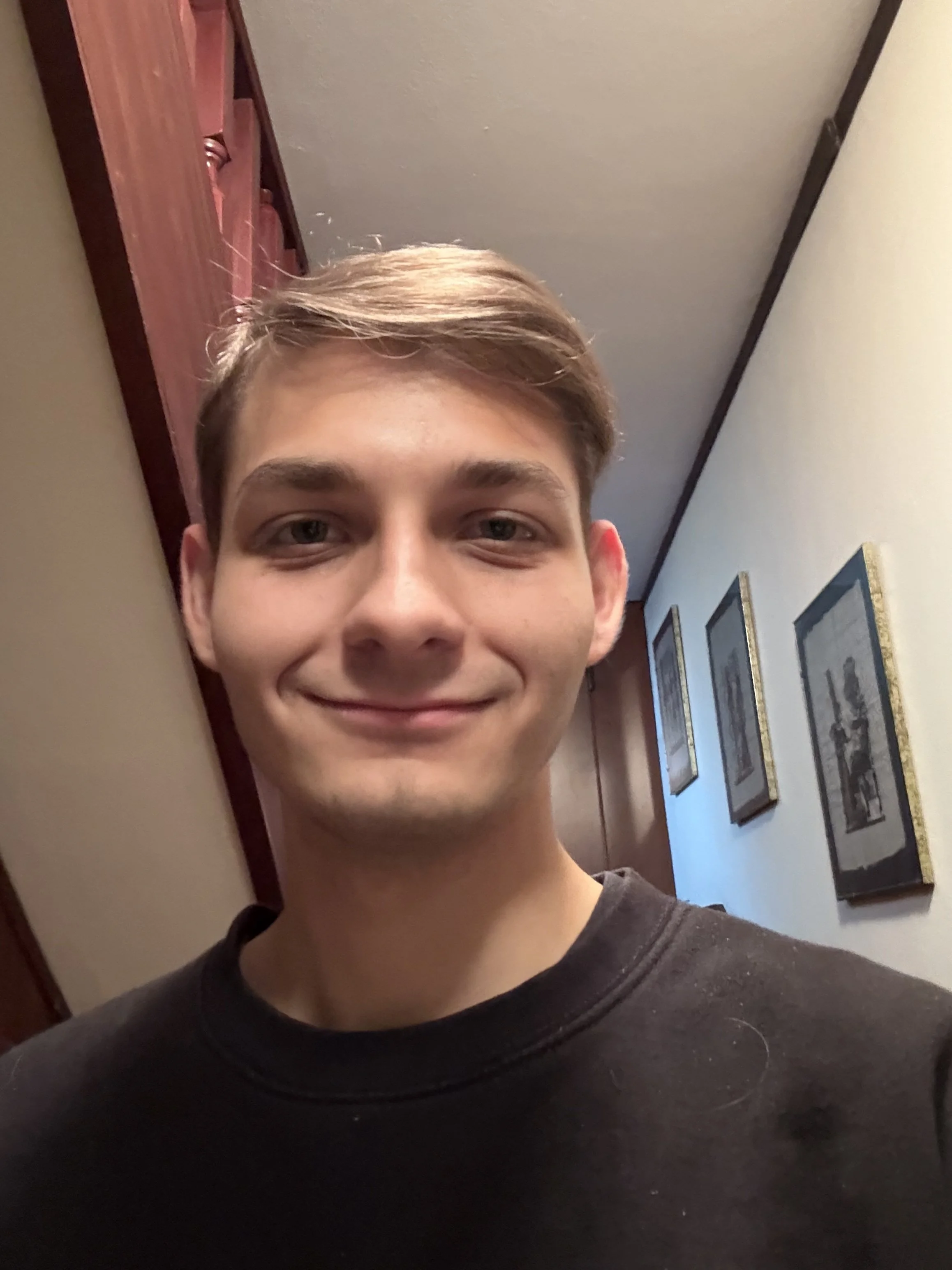 A young man with short brown hair smiling in a hallway with framed pictures on the wall.