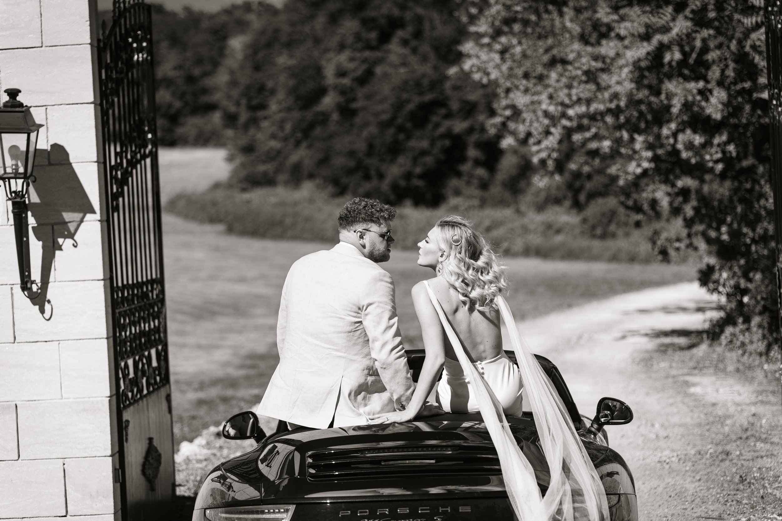 A black and white photo of a man and woman sitting on the back of a sports car, possibly a Porsche, near an open gate, with lush greenery in the background. The woman is wearing a backless dress with a long flowing train, and the man is dressed in a 