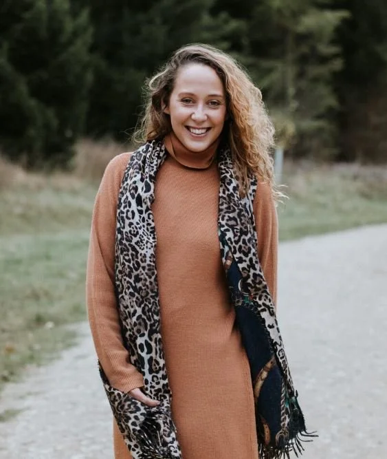 A smiling woman with curly hair wearing a brown sweater dress and a leopard print scarf standing outdoors on a gravel path with trees in the background.