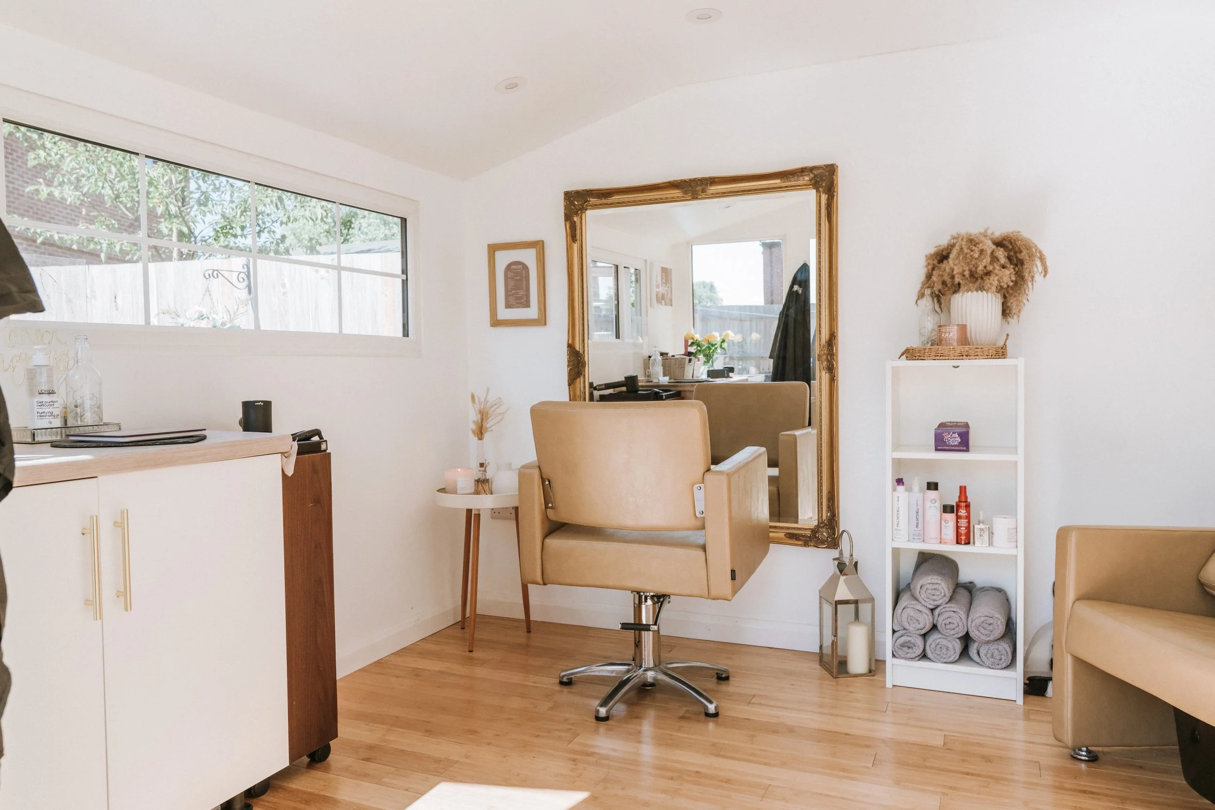 A salon in Bucks with a large gold-framed mirror, a beige salon chair, a small white shelf with rolled towels and beauty products, and a window letting in natural light.