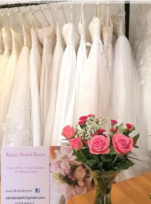A row of white wedding dresses hanging on a rack with lace and satin details, and a bouquet of pink roses in a glass vase on a wooden surface in front of the dresses.
