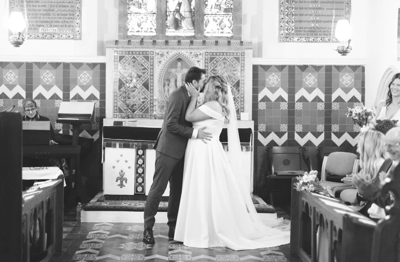 A couple shares a kiss during their wedding ceremony inside a church, with guests clapping and smiling, and the altar decorated with religious symbols and banners. Buckinghamshire