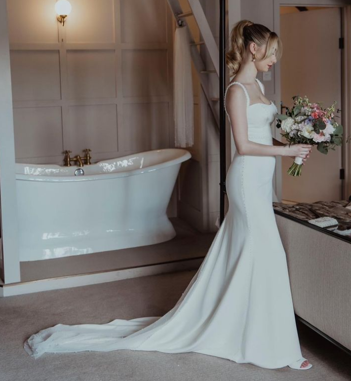 Bridal gown with train, bride holding bouquet, standing in a room near a mirror and bathroom with bathtub. The Kings Chapel, Amersham