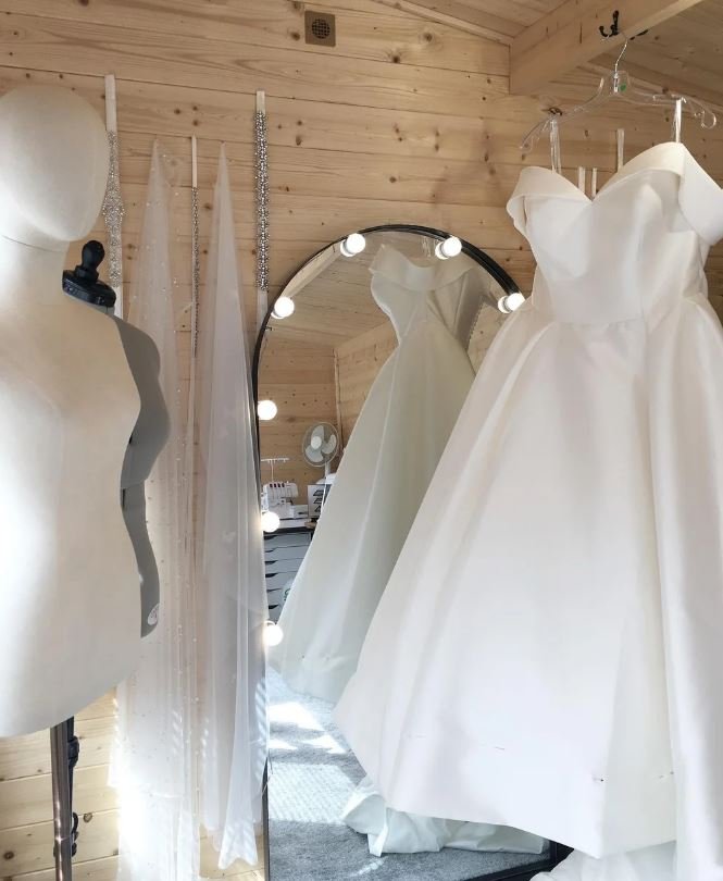 A fitting room with a mannequin, a wedding dress hanging on a hook, and a mirror decorated with string lights reflecting the wedding gown.