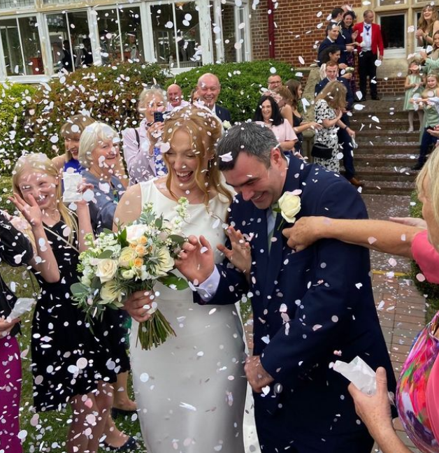 Bride and groom celebrate their wedding with confetti being thrown by guests outside a brick building, surrounded by friends and family.