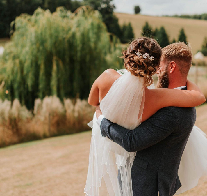 A newlywed couple embracing outdoors during daytime, with the groom holding the bride, in a scenic rural setting. Hadsham Farm, Oxfordshire
