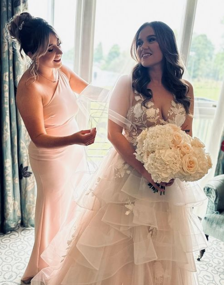 Two women, one in a wedding dress holding a bouquet of white roses, smiling at each other in a room with large windows and curtains. The Compleat Angler, Marlow