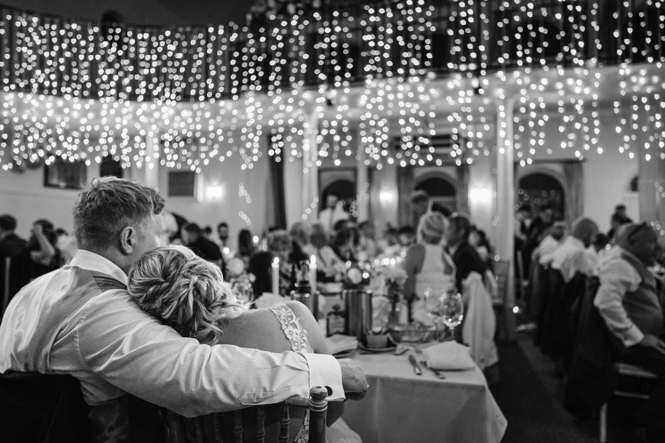 A black and white photo of a large indoor wedding reception with string lights hanging from the ceiling, many guests seated at decorated tables, and a couple sitting close together at the front. Kings Chapel, Amersham