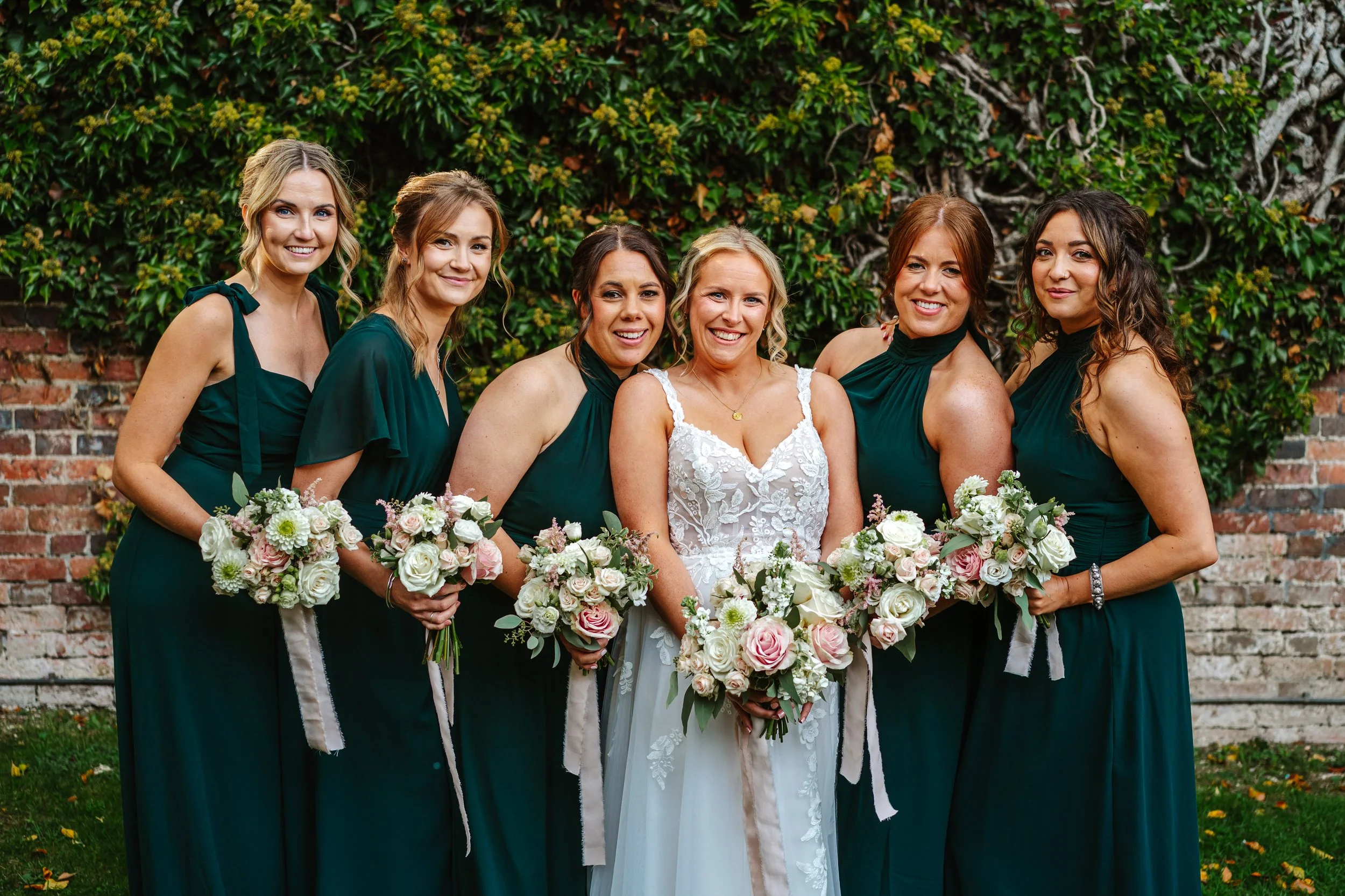 Group of six women in wedding and bridesmaid dresses holding floral bouquets standing outdoors in front of a brick and green vine-covered wall. Kings Chapel, Amersham