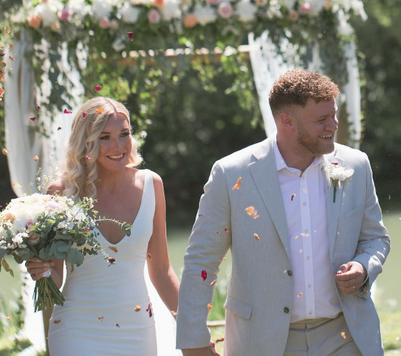 A bride and groom celebrate outdoors, smiling and surrounded by falling flower petals. The bride holds a bouquet, and the groom wears a light gray suit with a white boutonniere.