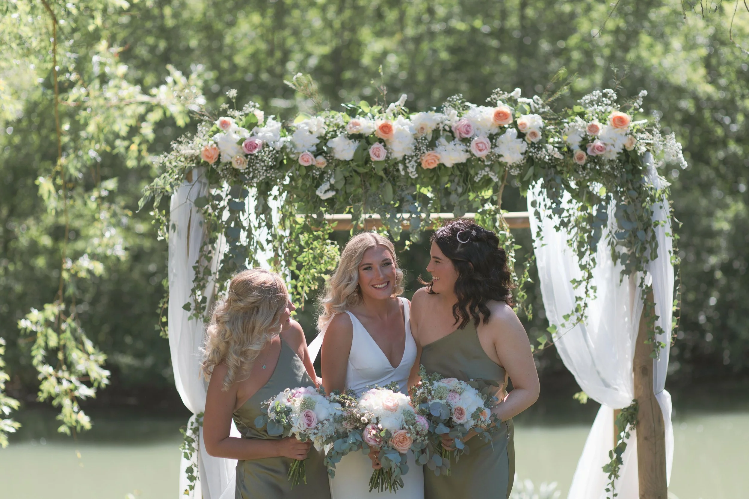 Three women in wedding attire standing under a floral arch outdoors, smiling and holding bouquets, with trees in the background. Bordeaux, France