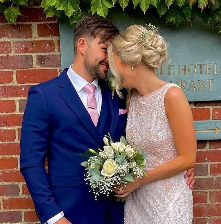 A newlywed couple on their wedding day, sharing a close moment with foreheads touching and eyes closed, standing in front of a brick wall with green foliage, the bride holding a bouquet of white roses and greenery. The Spread Eagle, Thame