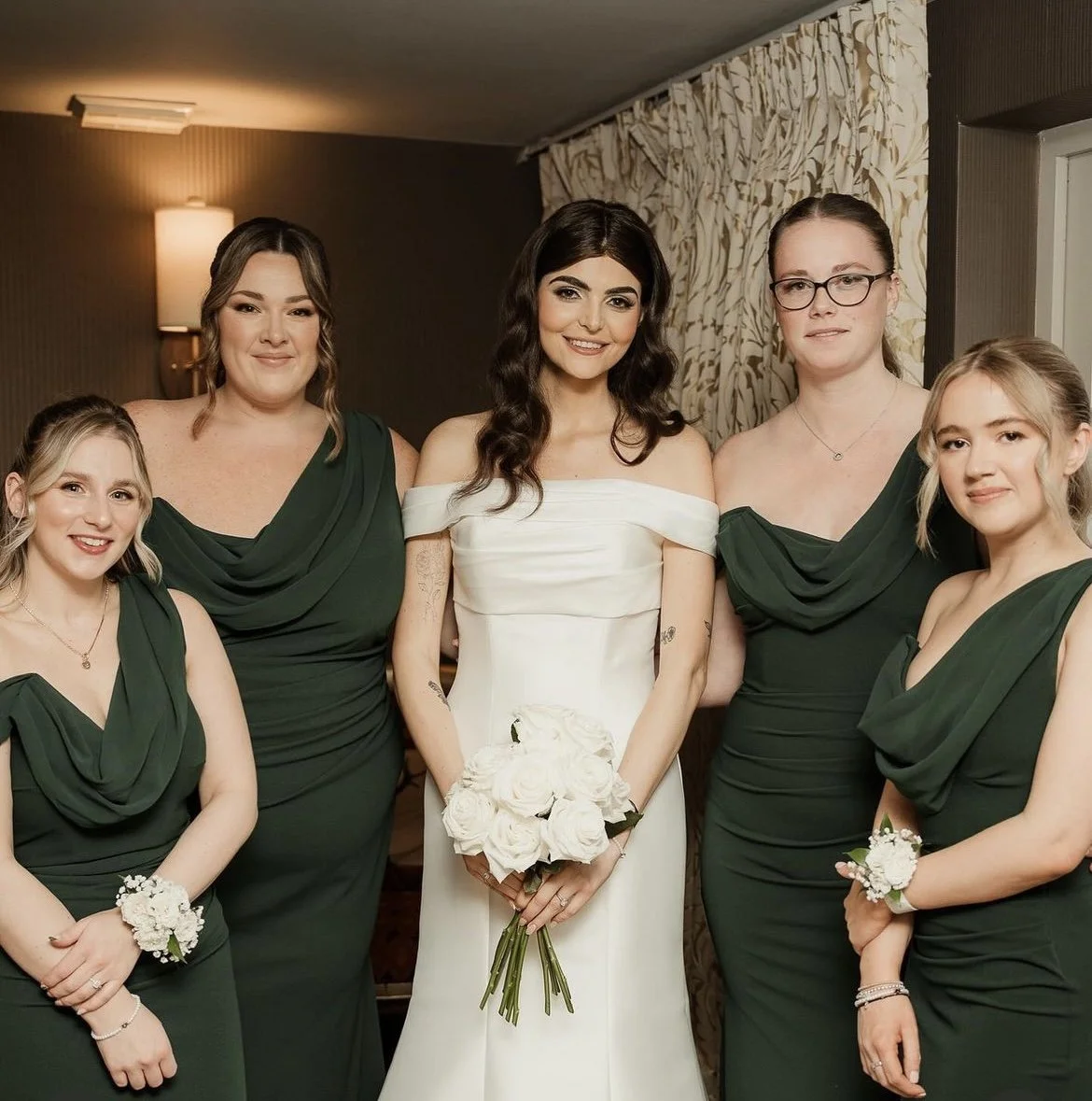 Group of women at a wedding, including the bride in a white dress holding a bouquet of white roses, and four bridesmaids in matching dark green dresses with floral wrist corsages, posing indoors. The Spread Eagle Thame