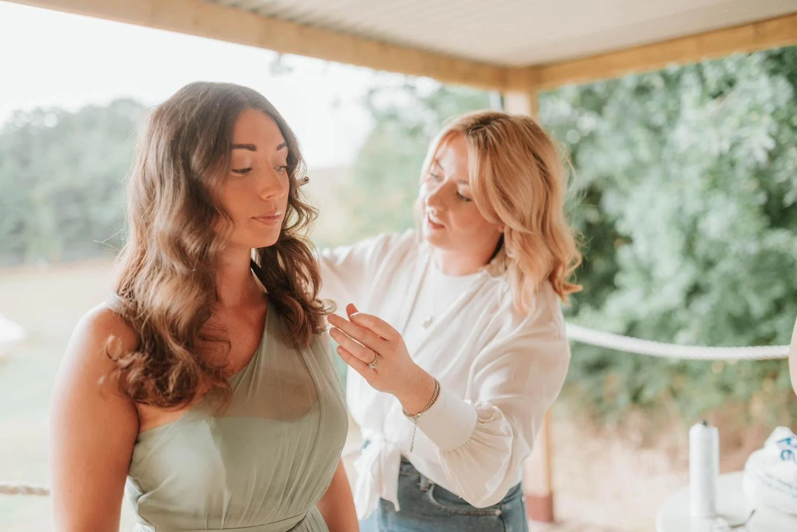 A woman in a beige dress is getting ready while another woman helps her with makeup or touch-up, outdoors with trees in the background.