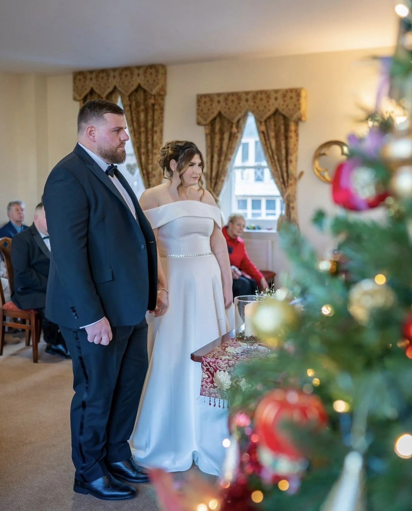 A bride and groom holding hands during their wedding ceremony, facing a decorated Christmas tree, with guests seated in the background in an ornately decorated room.