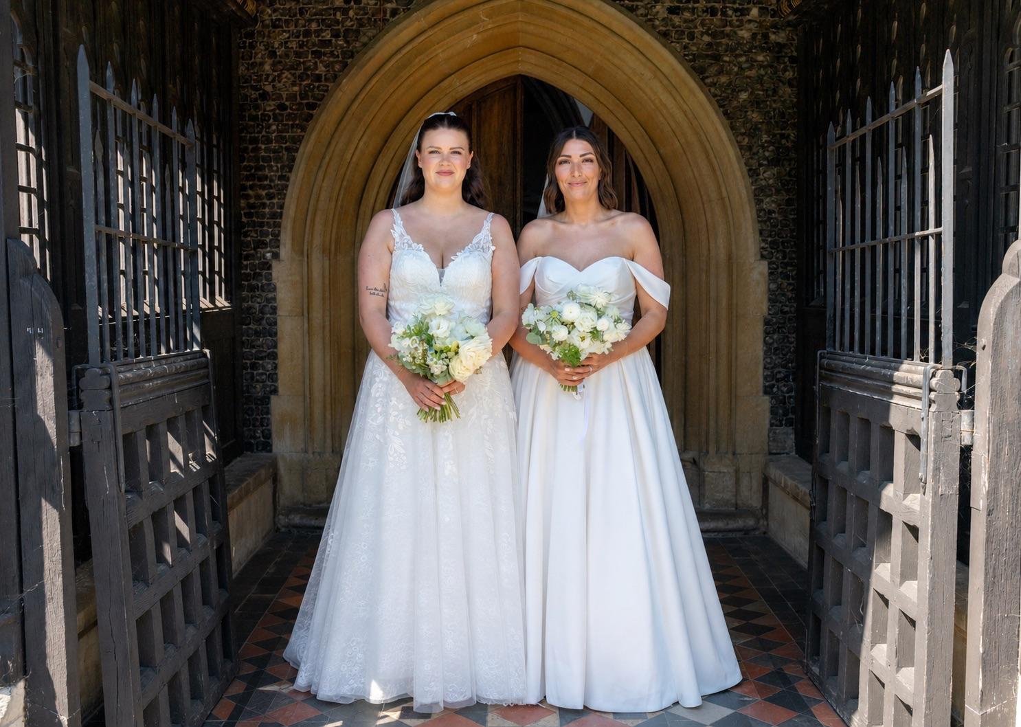 Two women in wedding dresses holding bouquets standing in front of a stone archway.