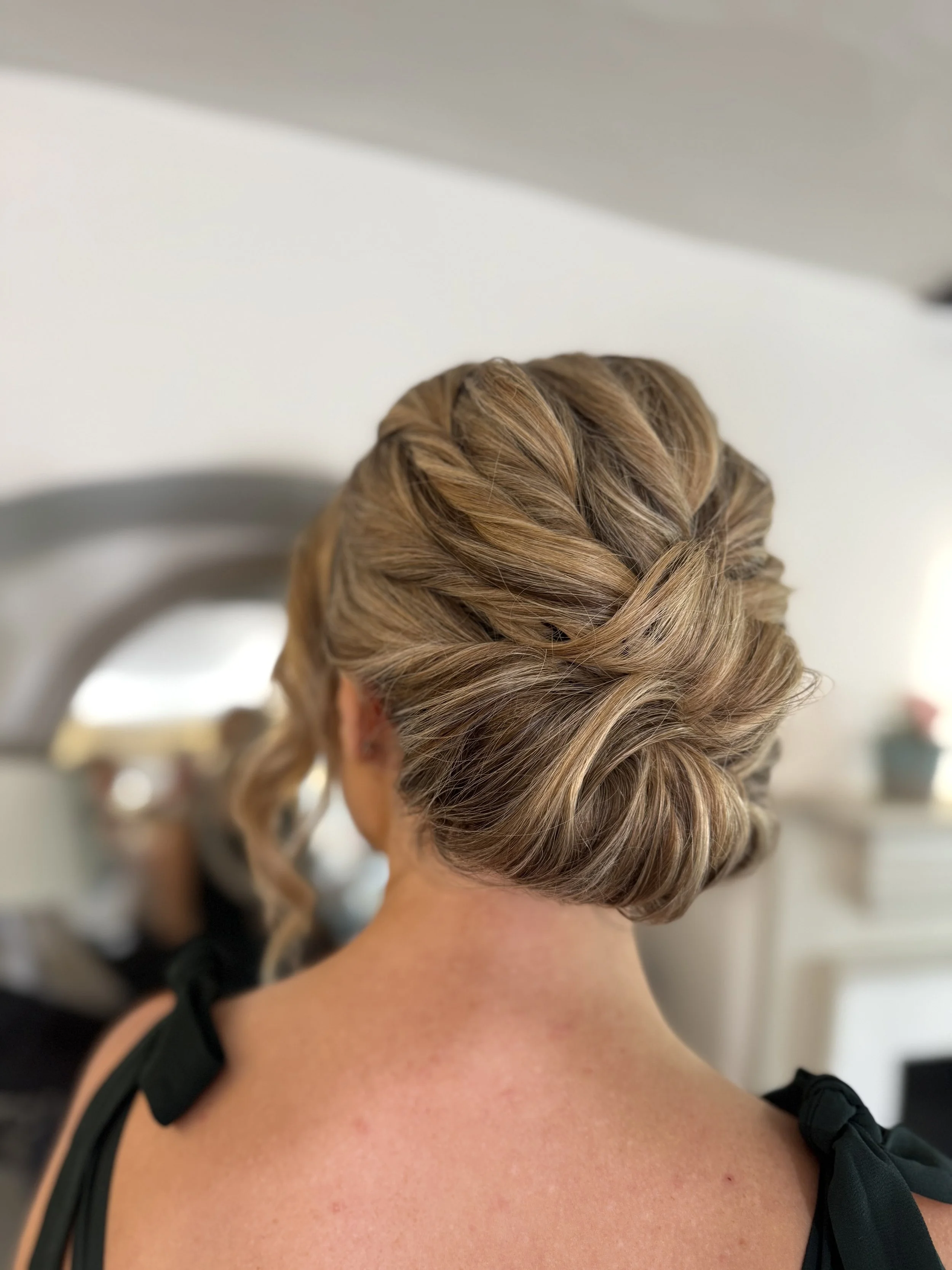 Back view of a woman with styled blonde hair in an elegant updo with twisted and braided sections, wearing a black dress with tied straps.