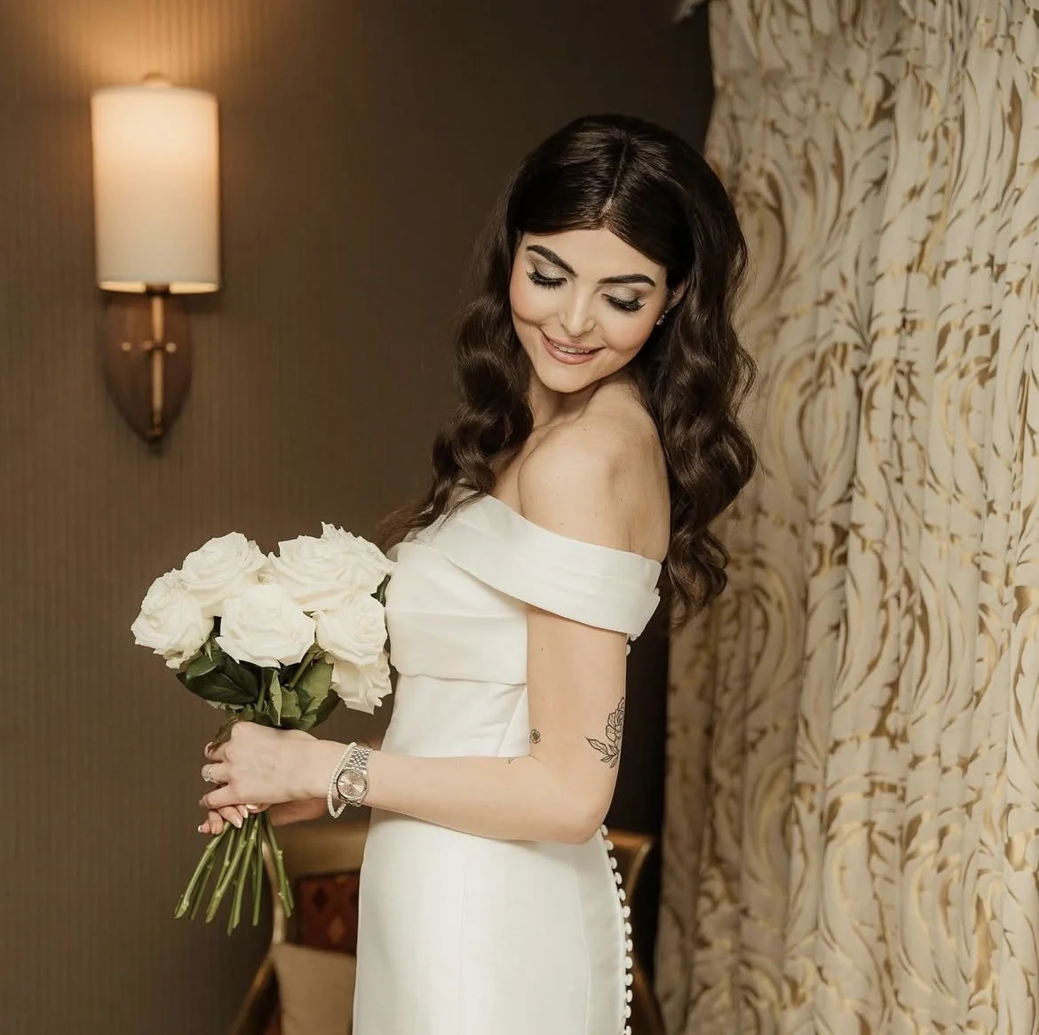 A young woman in a white off-shoulder dress holding a bouquet of white roses, smiling softly while looking down. The Spread Eagle Thame