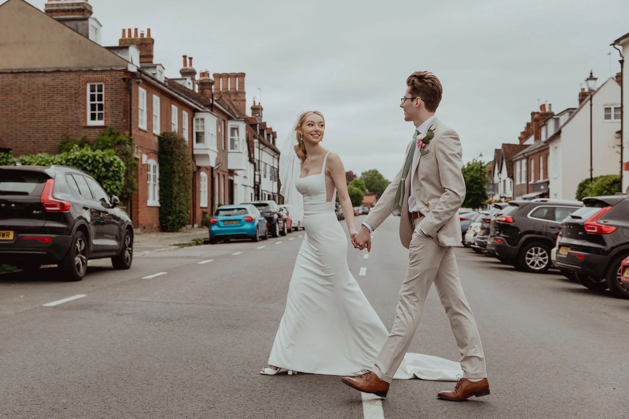 A couple in wedding attire holding hands and walking across a city street with parked cars and residential buildings in the background.