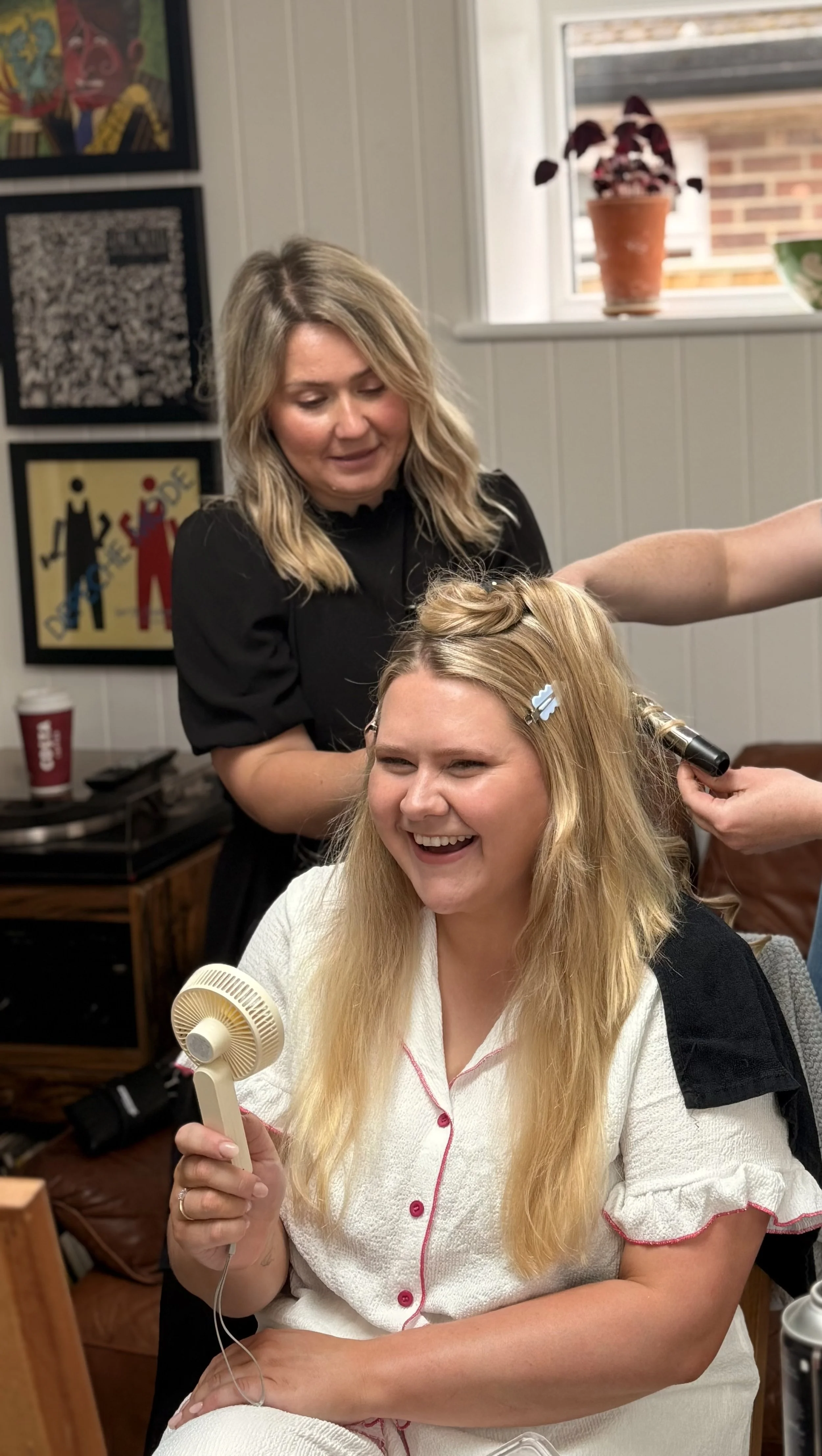 A woman with blonde hair, sitting and laughing, getting her hair styled by a hairdresser. The hairdresser is standing behind her, using an iron on her hair, while the woman holds a small handheld fan and a curling iron.