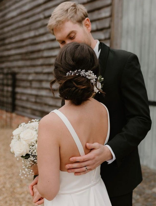 A bride and groom embrace outdoors, with the groom in a black tuxedo and the bride holding a bouquet of white flowers, against a rustic wooden background.