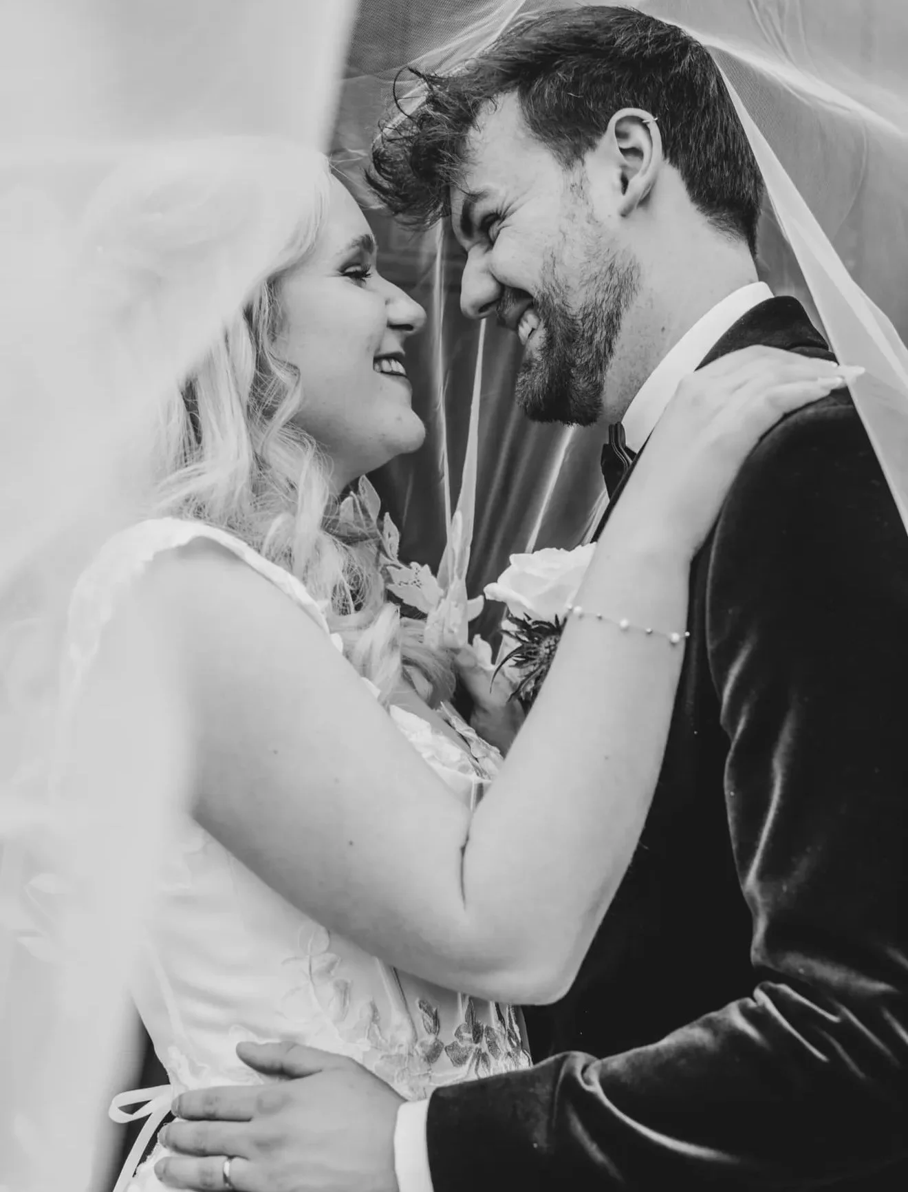 Black and white photograph of a bride and groom smiling at each other, with the bride wearing a veil and the groom in a tuxedo. They are close, with their foreheads touching.