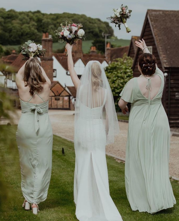 A bride and two bridesmaids are seen from behind, celebrating outdoors with bouquets raised, in a rustic village setting.