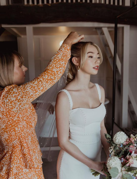 Bride getting her veil adjusted by a woman in a orange patterned dress, holding a bouquet of flowers.