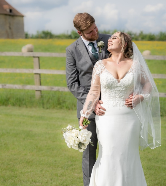 Bride and groom on a grassy field during their wedding, with a wooden fence and a building in the background.