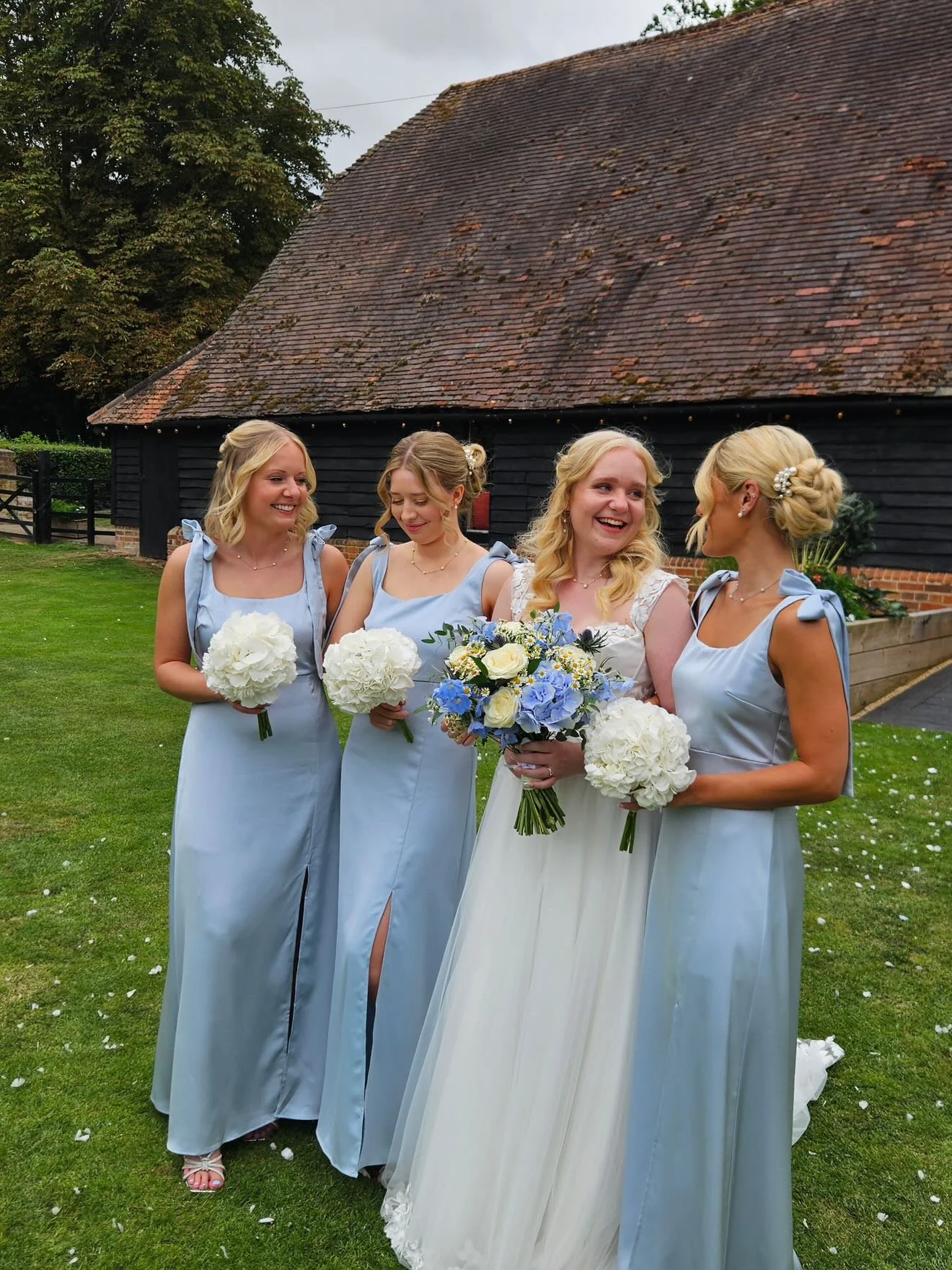 A group of four women, including a bride and her bridesmaids, standing on a lawn with a rustic building in the background. They are dressed in elegant dresses, holding bouquets, and smiling. Oxfordshire